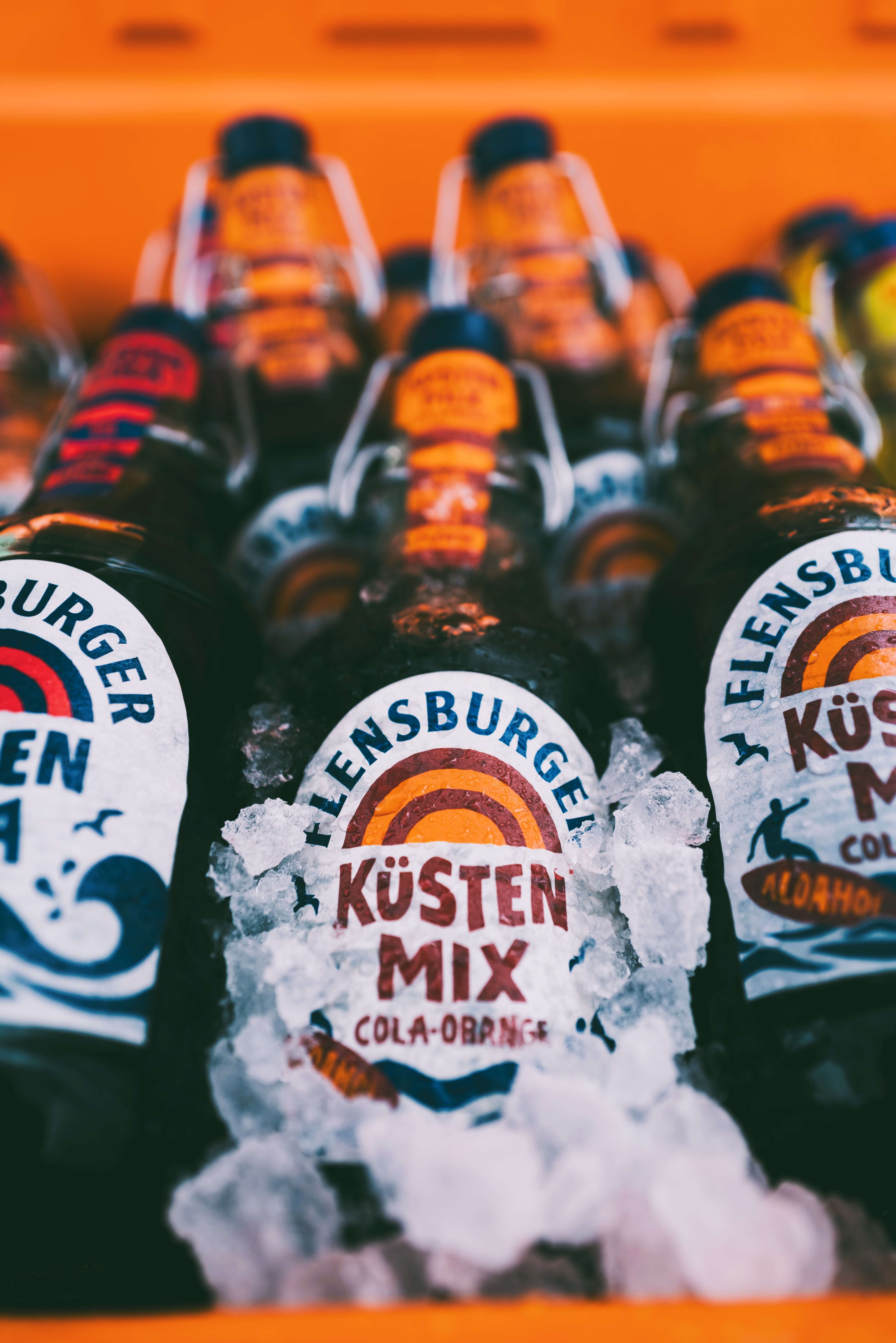 Flensburger Küstenmix "Cola-Orange" Flaschen liegen dekorativ auf Eis. Die Etiketten zeigen den Namen, den Alkoholgehalt von 0,0% und die Menge von 0,33 Litern an.