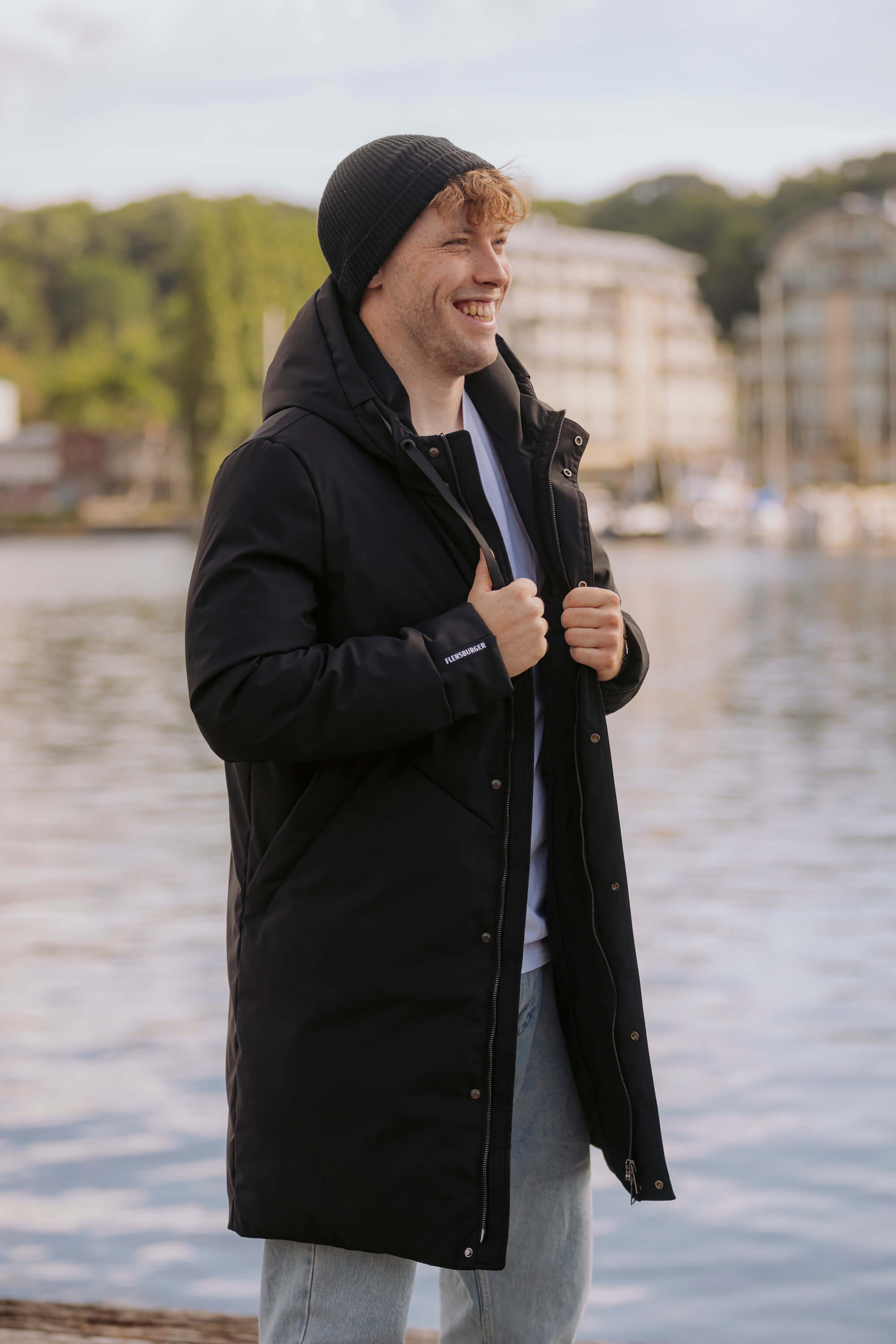 Mann trägt einen schwarzen FLENS Winterparka. Er lächelt und hält den Parka am Reißverschluss, im Hintergrund ist Wasser mit Booten und Gebäuden zu sehen.