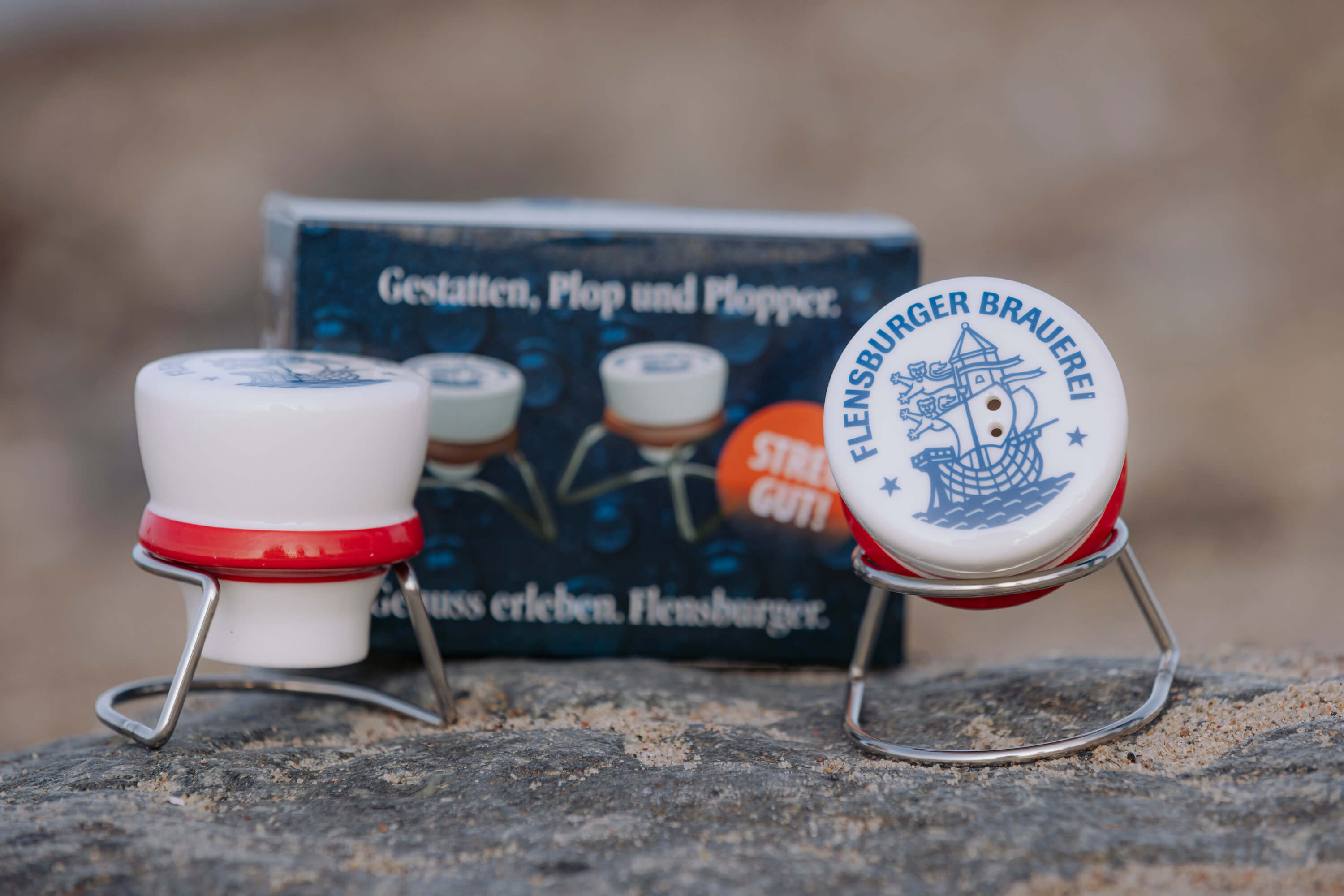 Zwei keramk Salz- und Pfefferstreuer mit Metallhalterung in Bügelverschlussoptik stehen auf einem Stein. Einer zeigt den Schriftzug „FLENSBURGER BRAUEREI“ und ein Schiff. Im Hintergrund ist eine blaue Verpackung mit weißer Schrift.