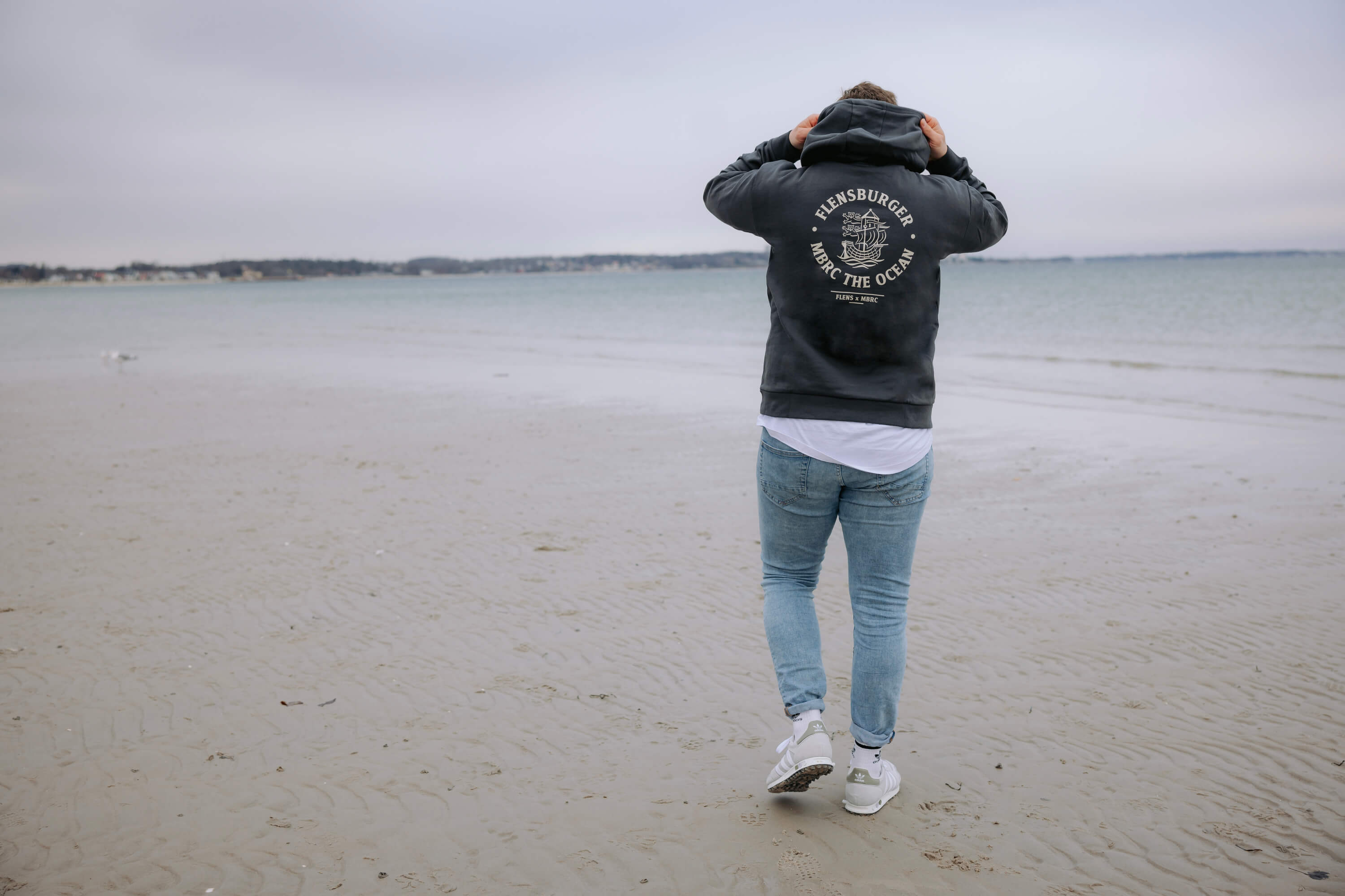Mann trägt Hoodie in Anthrazit mit Aufdruck "FLENSBURGER MBRC THE OCEAN" auf dem Rücken am Strand. Er zieht die Kapuze über den Kopf und blickt aufs Wasser.