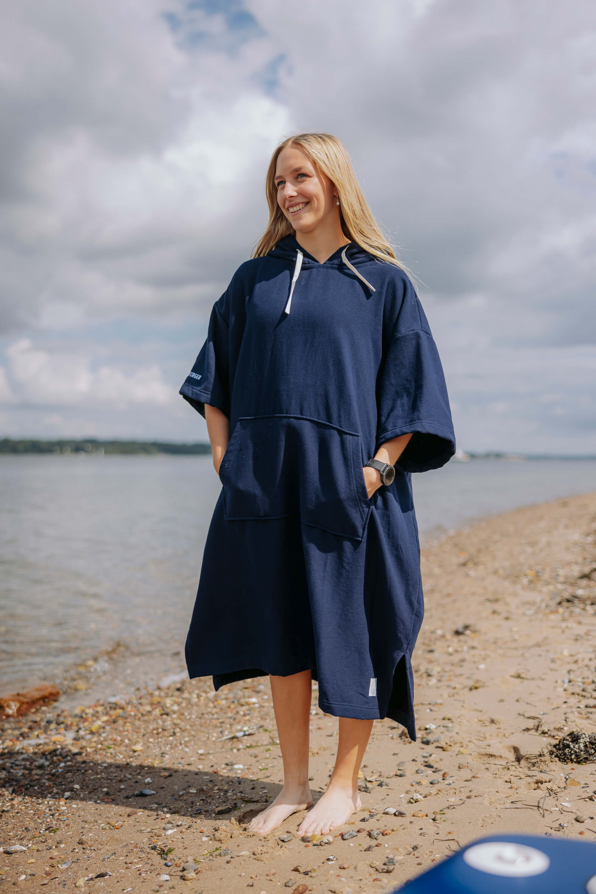 Frau trägt einen blauen Strand-Poncho mit Kapuze und kurzen Ärmeln. Sie steht barfuß am Strand und lächelt bei bewölktem Himmel.
