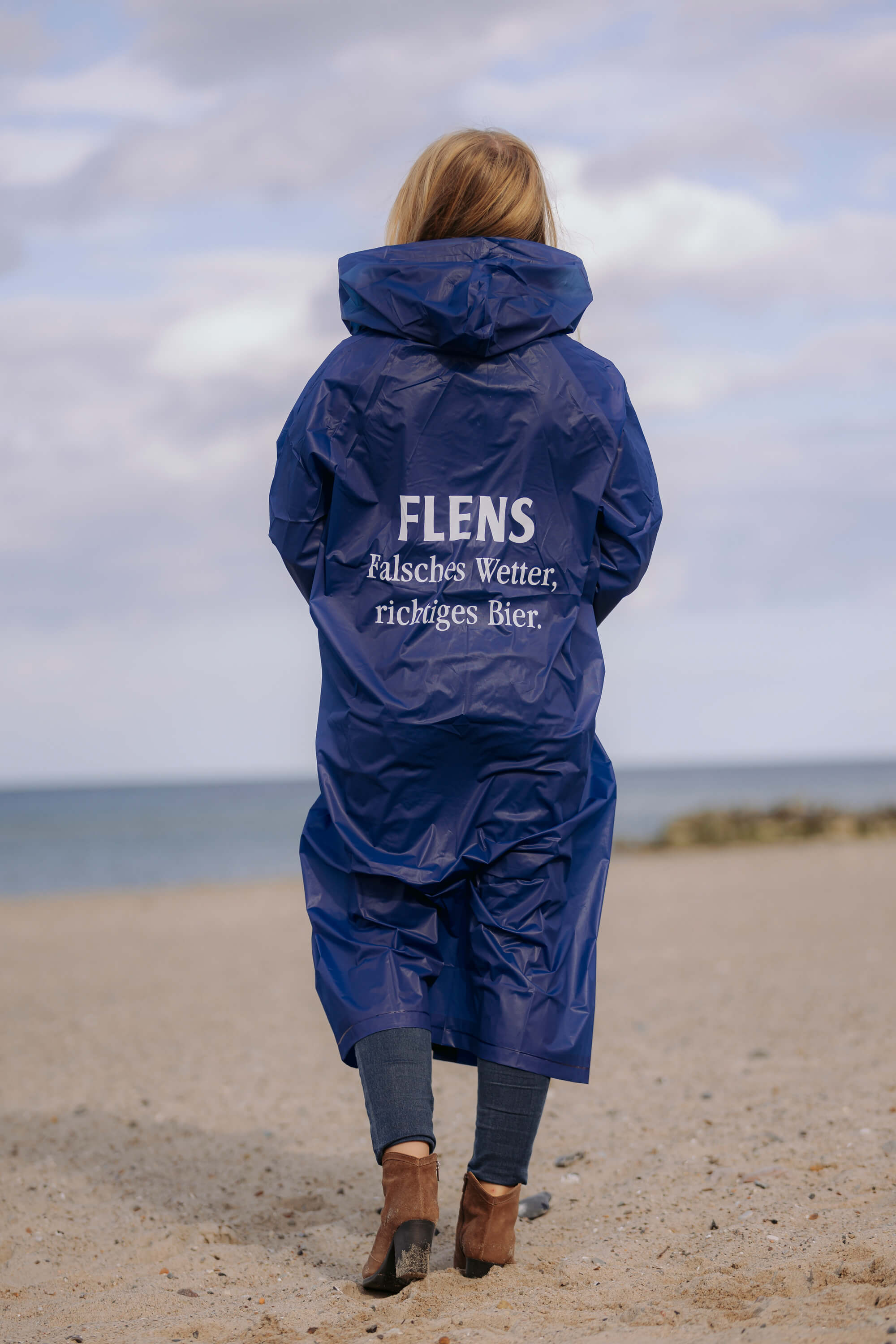 Person steht mit dem Rücken zur Kamera und trägt einen blauen Regenponcho mit Kapuze. Auf dem Rücken steht in weißer Schrift: „FLENS Falsches Wetter, richtiges Bier.“ Person steht am Strand, Blick aufs Meer.