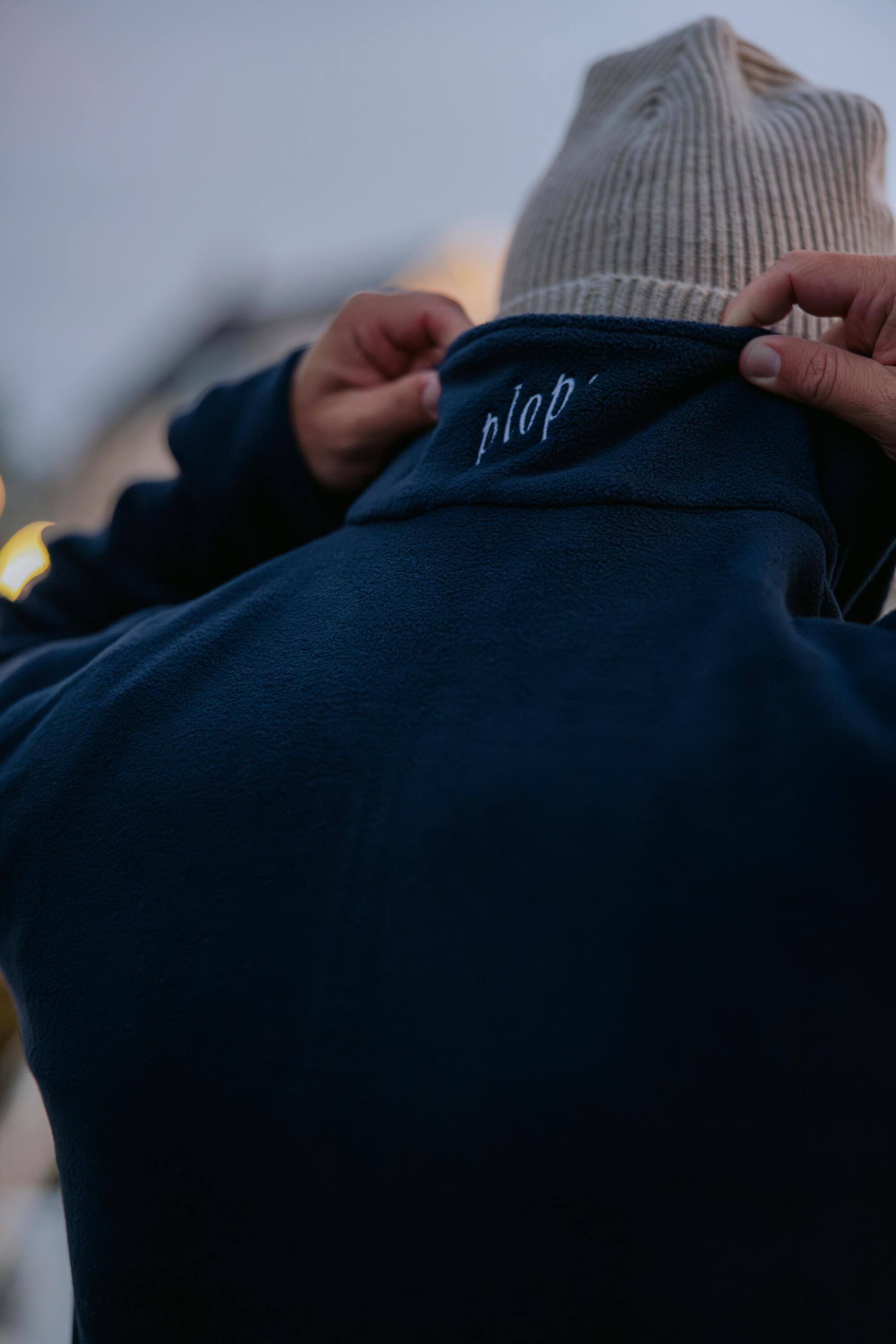 Person zieht den Kragen eines dunkelblauen Fleece-Pullovers mit gesticktem Schriftzug "plop" am Kragen hoch. Person trägt eine hellgraue Strickmütze.