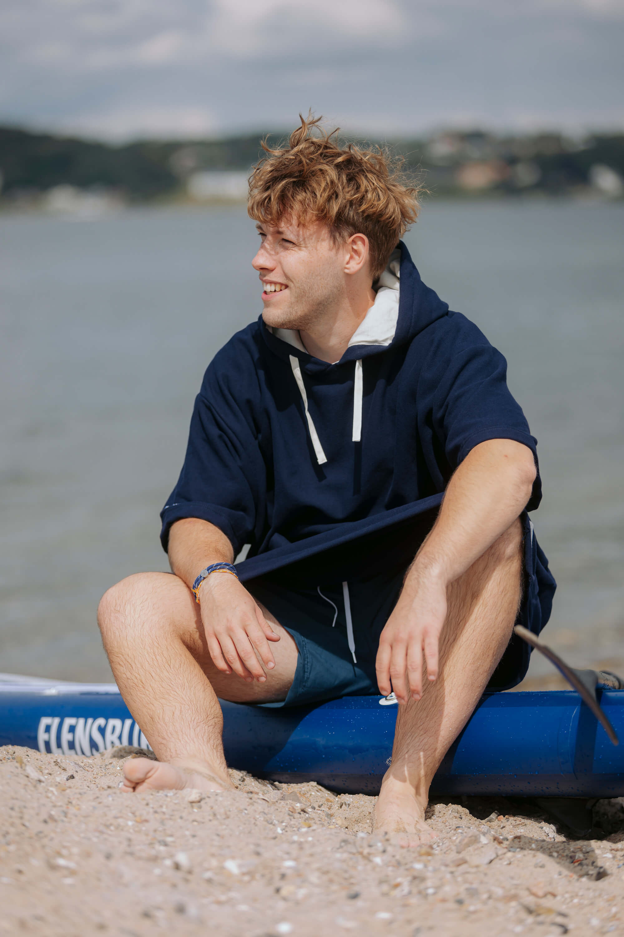 Junger Mann sitzt barfuß auf einem dunkelblauen SUP-Board am Strand und trägt einen blauen Strand-Poncho mit weißen Kordeln.