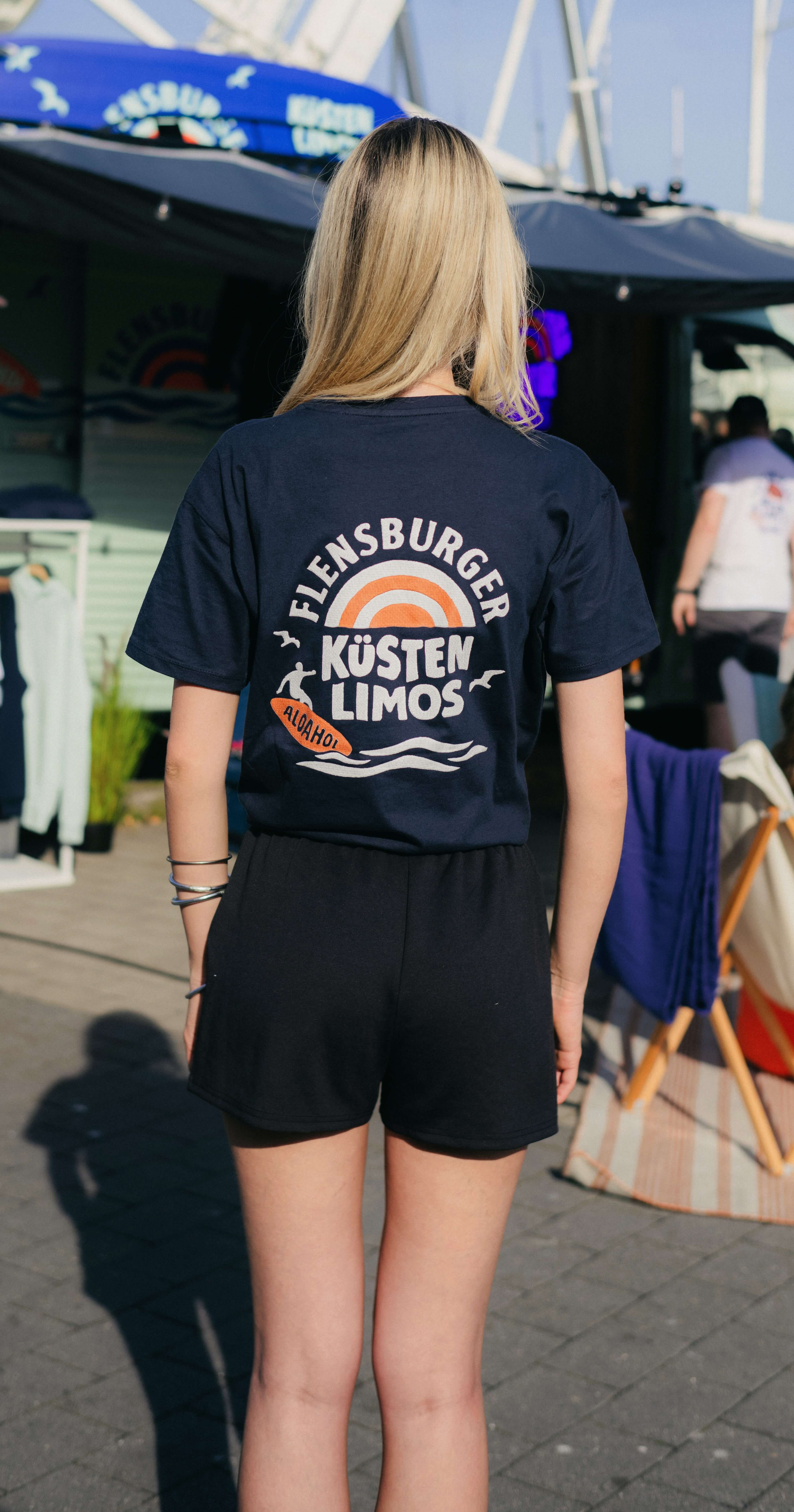 Junge Frau trägt ein navyblaues T-Shirt mit dem Aufdruck „FLENSBURGER KÜSTEN LIMOS" und steht vor einem Verkaufsstand. Sie trägt schwarze Shorts und hat lange, blonde Haare.