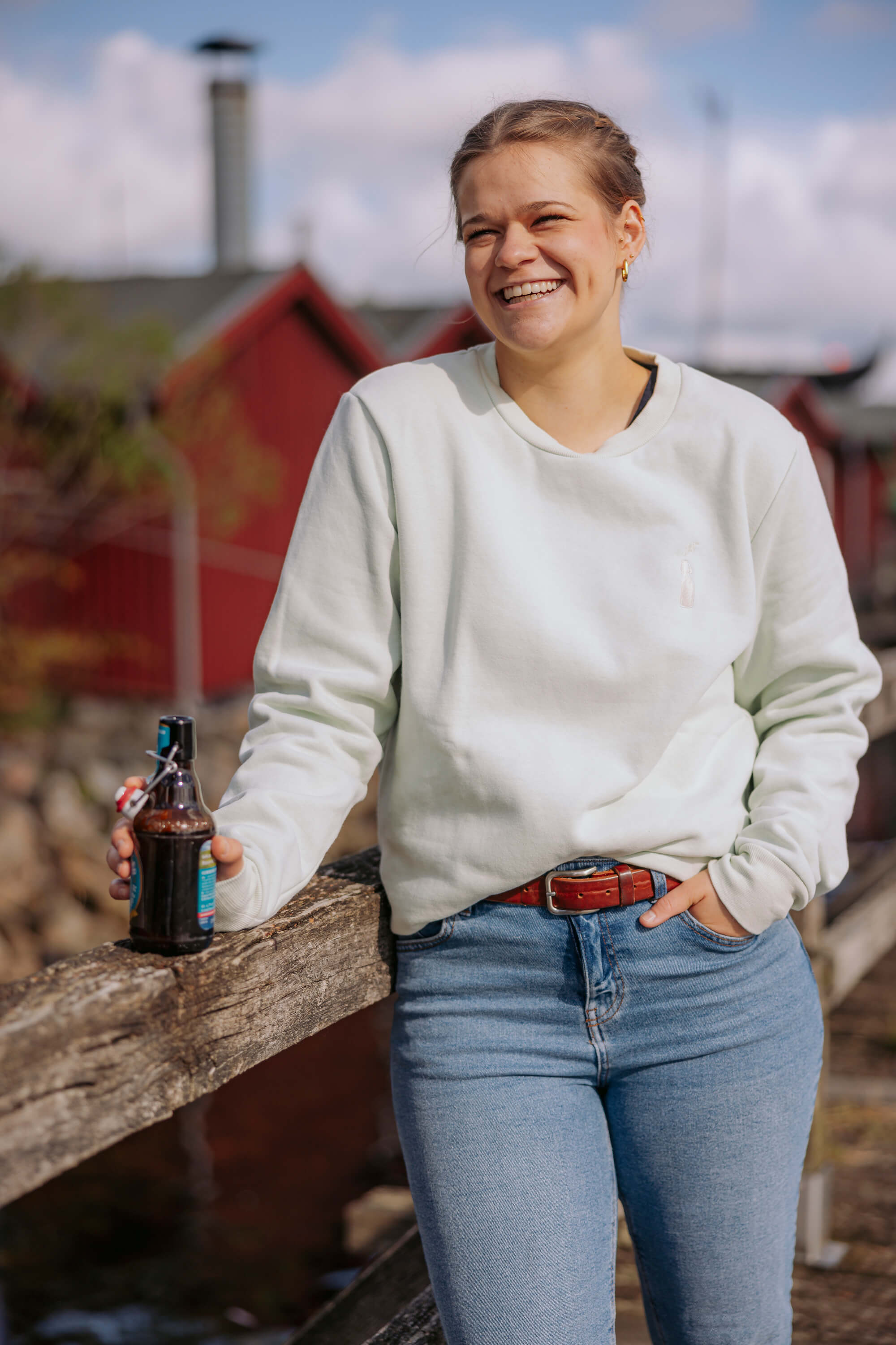 Junge Frau trägt weißen Sweater "Sommerbrise" und hellblaue Jeans. Sie lehnt an Holzbalken und hält eine braune Bügelflasche in der linken Hand.