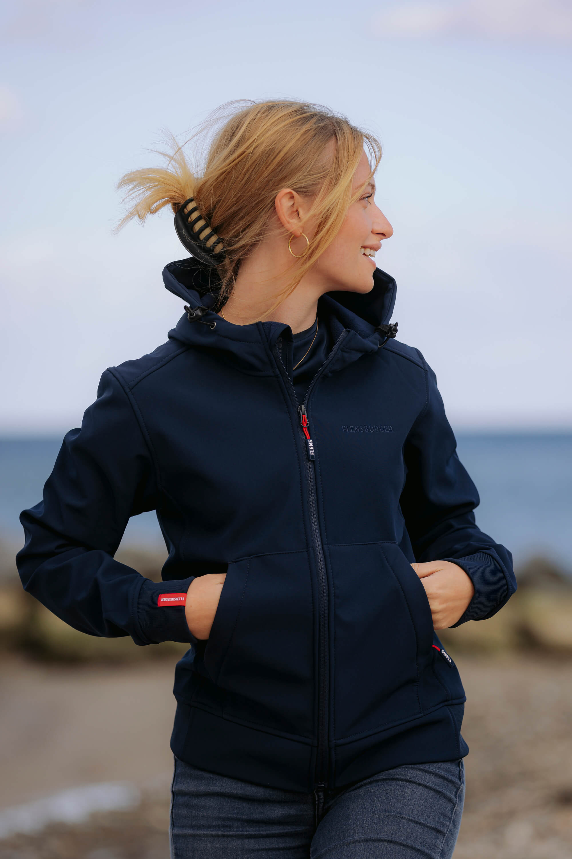 Frau trägt eine navyblaue Softshelljacke mit Kapuze und Reißverschluss. Sie hat beide Hände in den Taschen und schaut lächelnd seitlich weg. Im Hintergrund ist das Meer zu sehen.