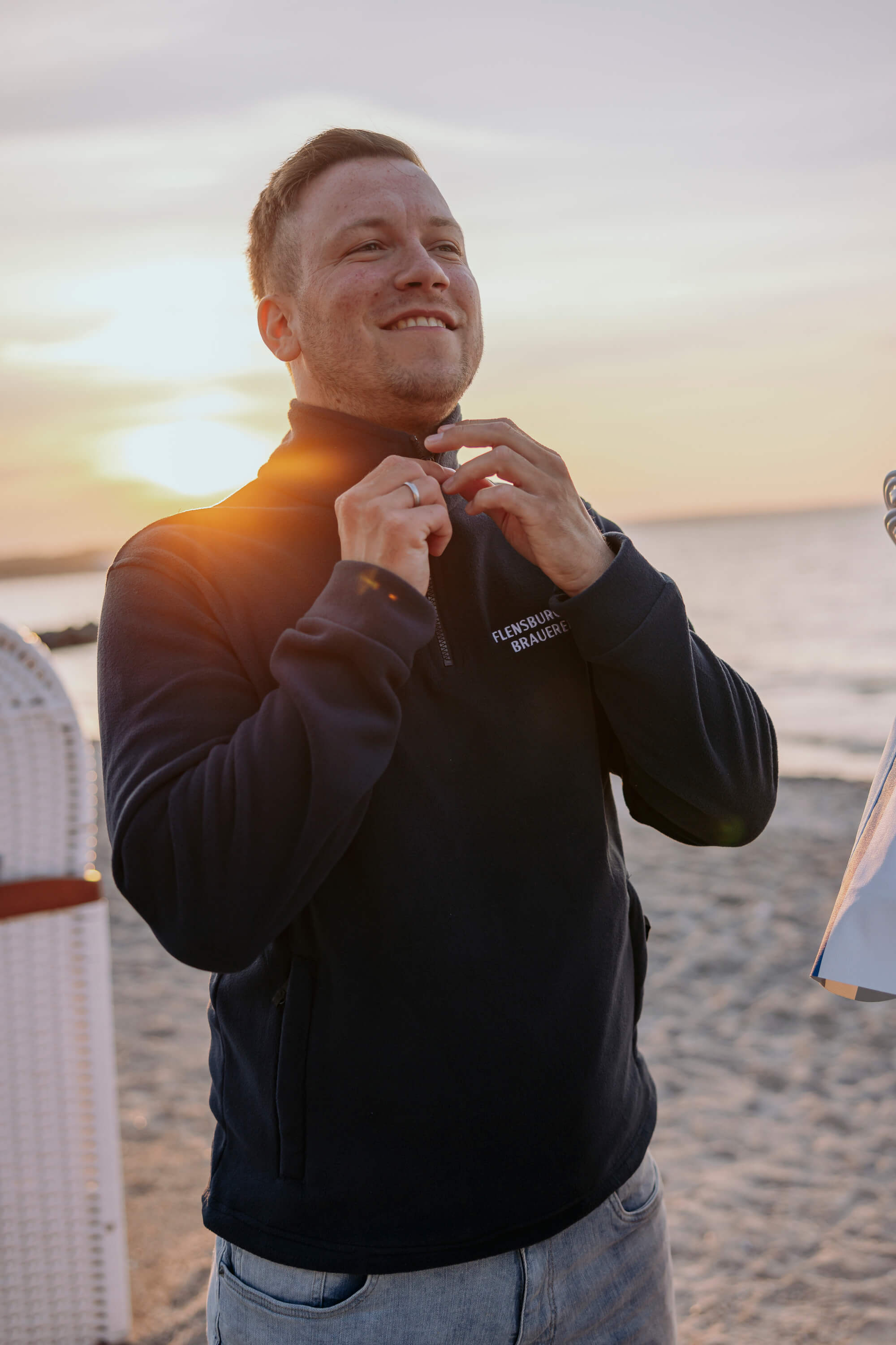 Mann steht am Strand bei Sonnenuntergang und schließt den Reißverschluss eines navyblauen Fleece-Pullovers mit Aufschrift „Flensburger Brauerei“.