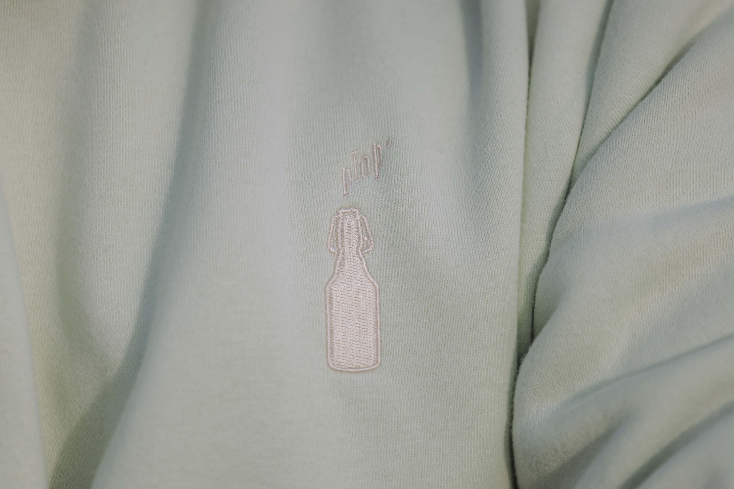 Datailsansicht des Brustbereichs eines Sweater. Auf dem mintgrünen Stoff ist eine weiße Stickerei einer Flasche mit der Schrift „plop'“ oberhalb angebracht.
