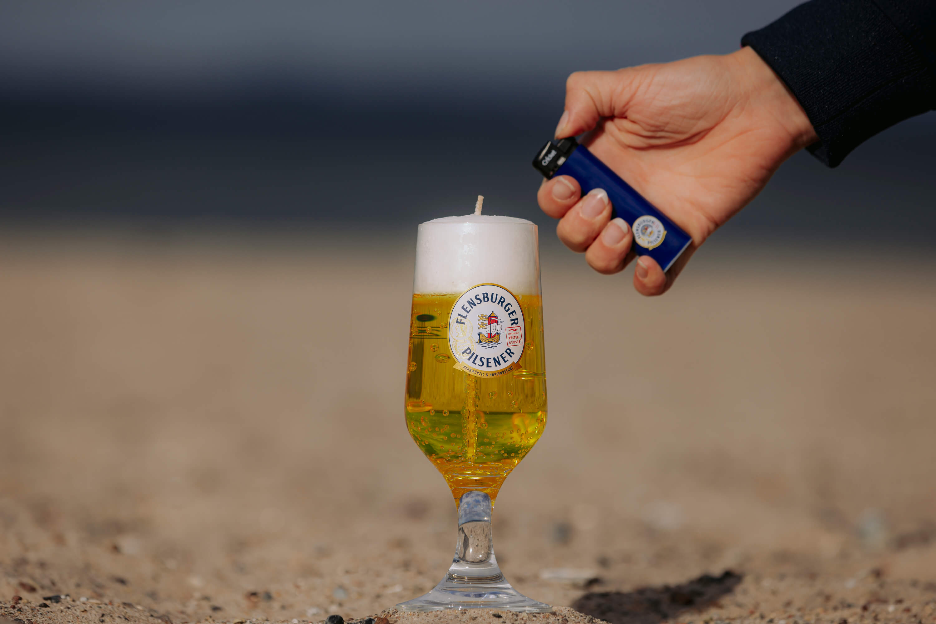 Hand hält blauen Feuerzeug und zündet Kerze in einem Flensburger Pilsener Glas an. Das Glas ist mit goldenem und weißem Wachs, um Bier zu immitieren.