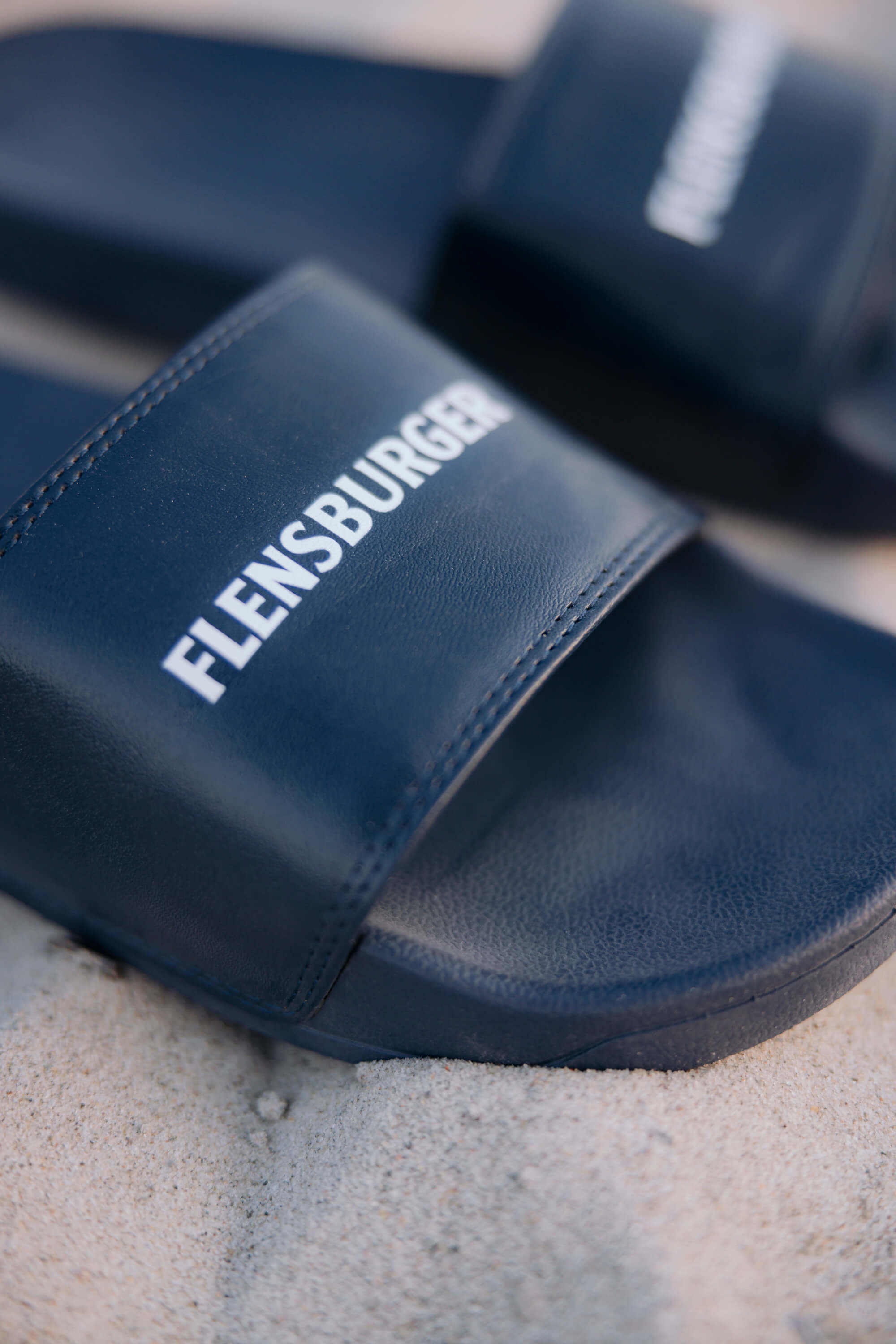 Dunkelblaue Fördeslipper mit weißer Aufschrift „FLENSBURGER“ liegen auf hellem Sand.