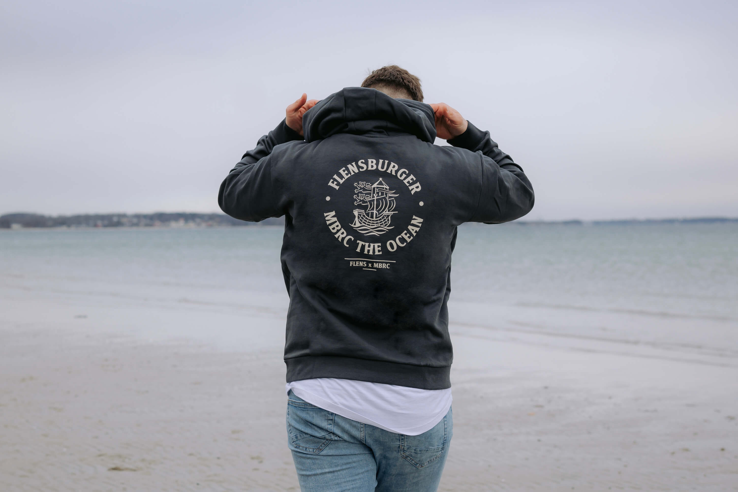 Person steht am Strand und zieht die Kapuze eines dunkelblauen Hoodies mit Aufdruck "FLENSBURGER MBRC THE OCEAN FLENS x MBRC" auf dem Rücken über den Kopf.
