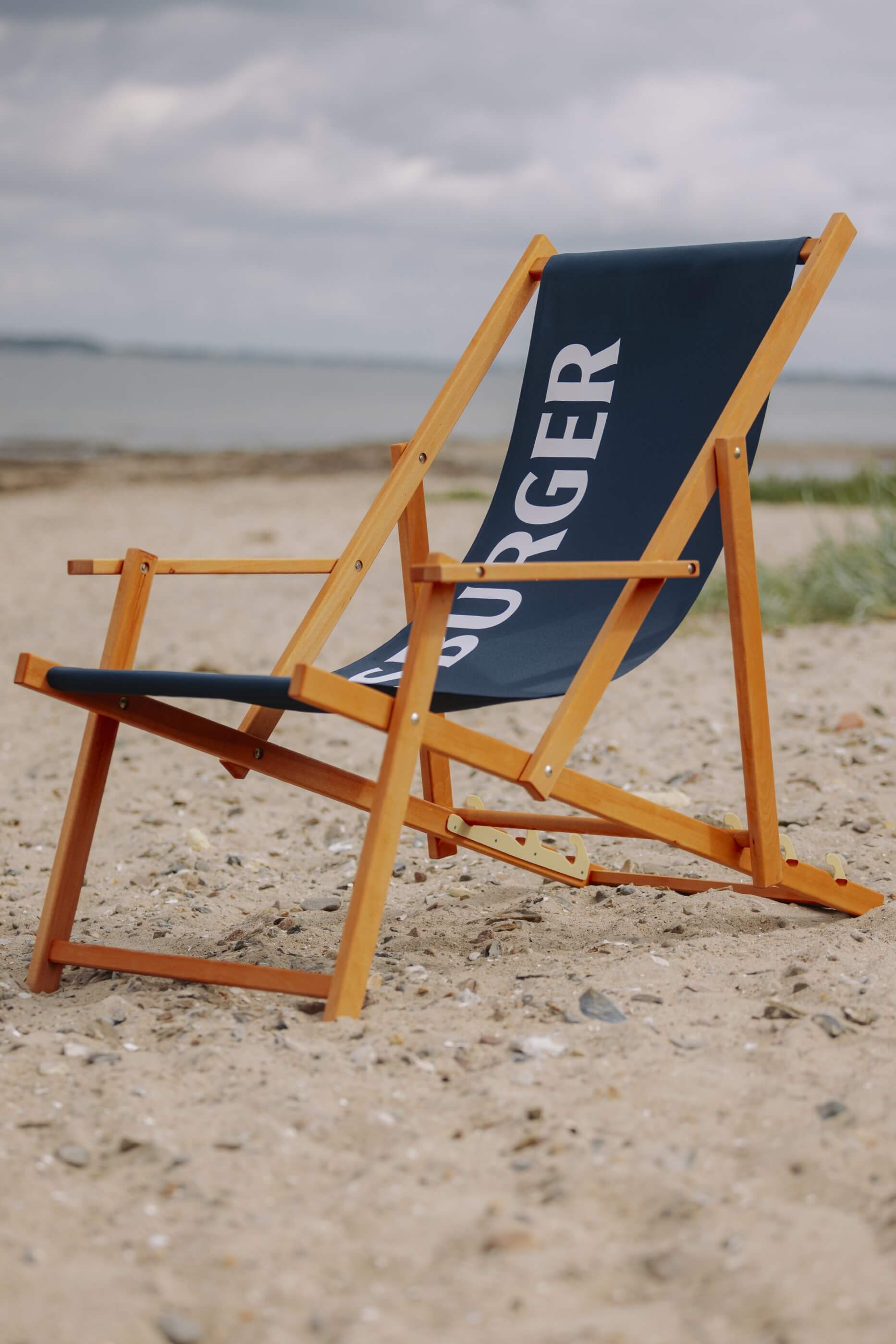 Liegestuhl aus hellem Buchenholz mit blauem Stoff, auf dem in großen weißen Buchstaben „FLENSBURGER“ steht, steht am sandigen Strand mit unscharfem Meer im Hintergrund.