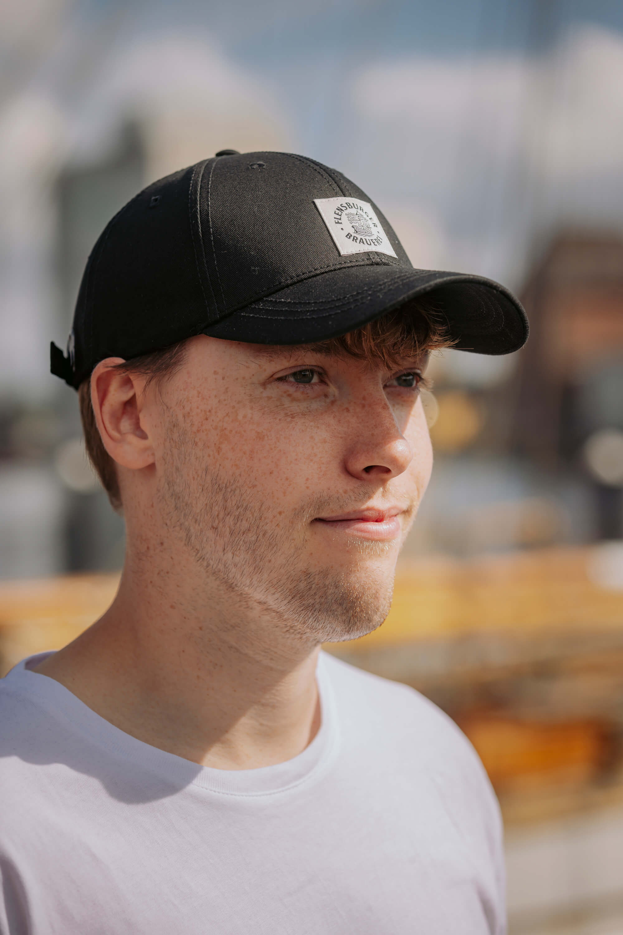 Junger Mann trägt eine schwarze Basic Cap mit einem weißen Weblabel, auf dem "FLENS BURGER BRAUEREI" und ein Brauereilogo zu sehen sind. Er trägt ein weißes T-Shirt und schaut leicht nach rechts.