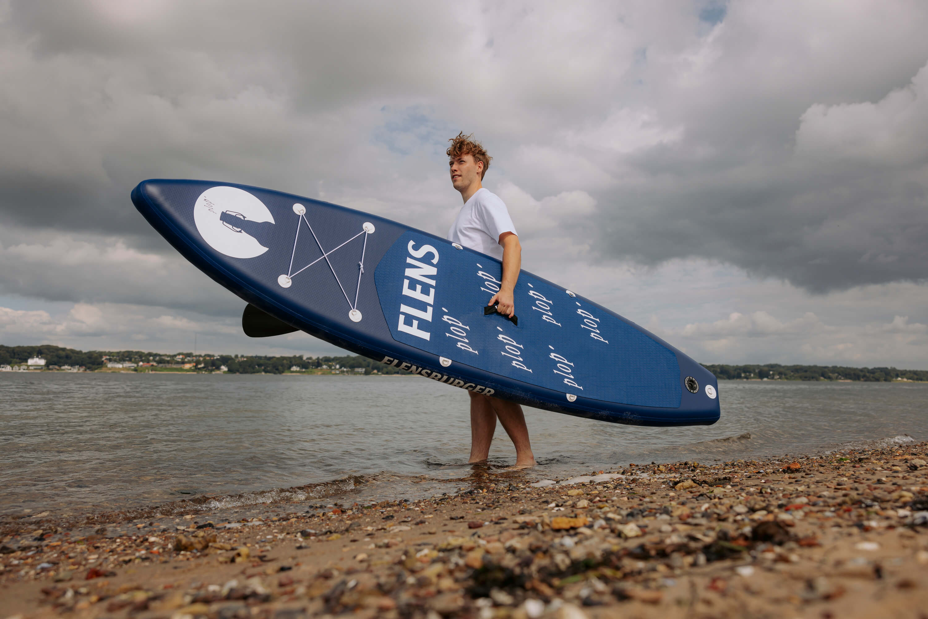 Junger Mann trägt das blaue FLENS SUP Board mit weißen Schriftzügen "FLENS" und "plop'" am Strand. Er steht im flachen Wasser unter bewölktem Himmel.