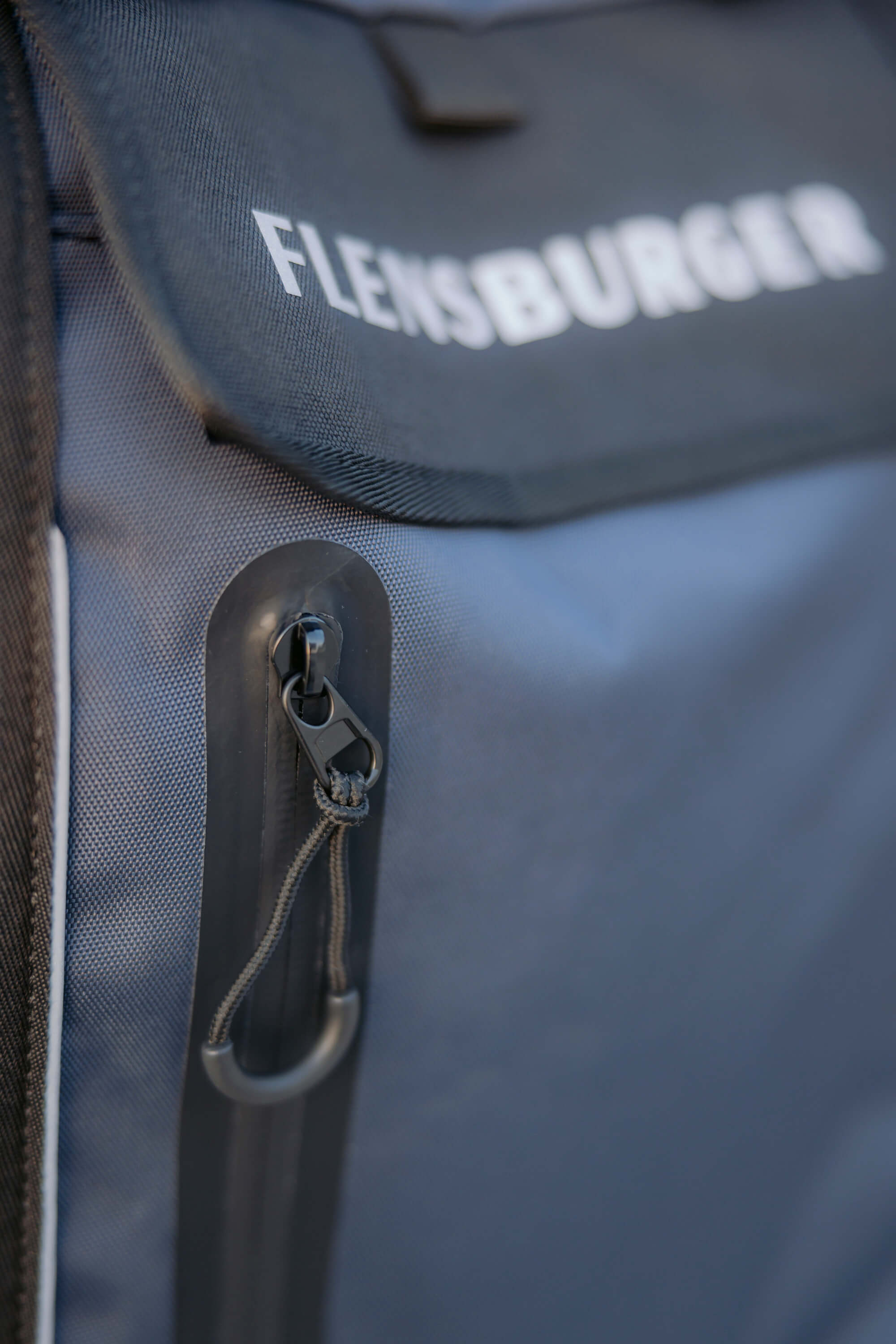Detailansicht eines dunkelblauen Rucksacks mit dem weißen Aufdruck „Flensburger“ auf der vorderen Klappe. Der Rucksack besitzt einen schwarzen Reißverschluss mit Zugband am vorderen Fach.