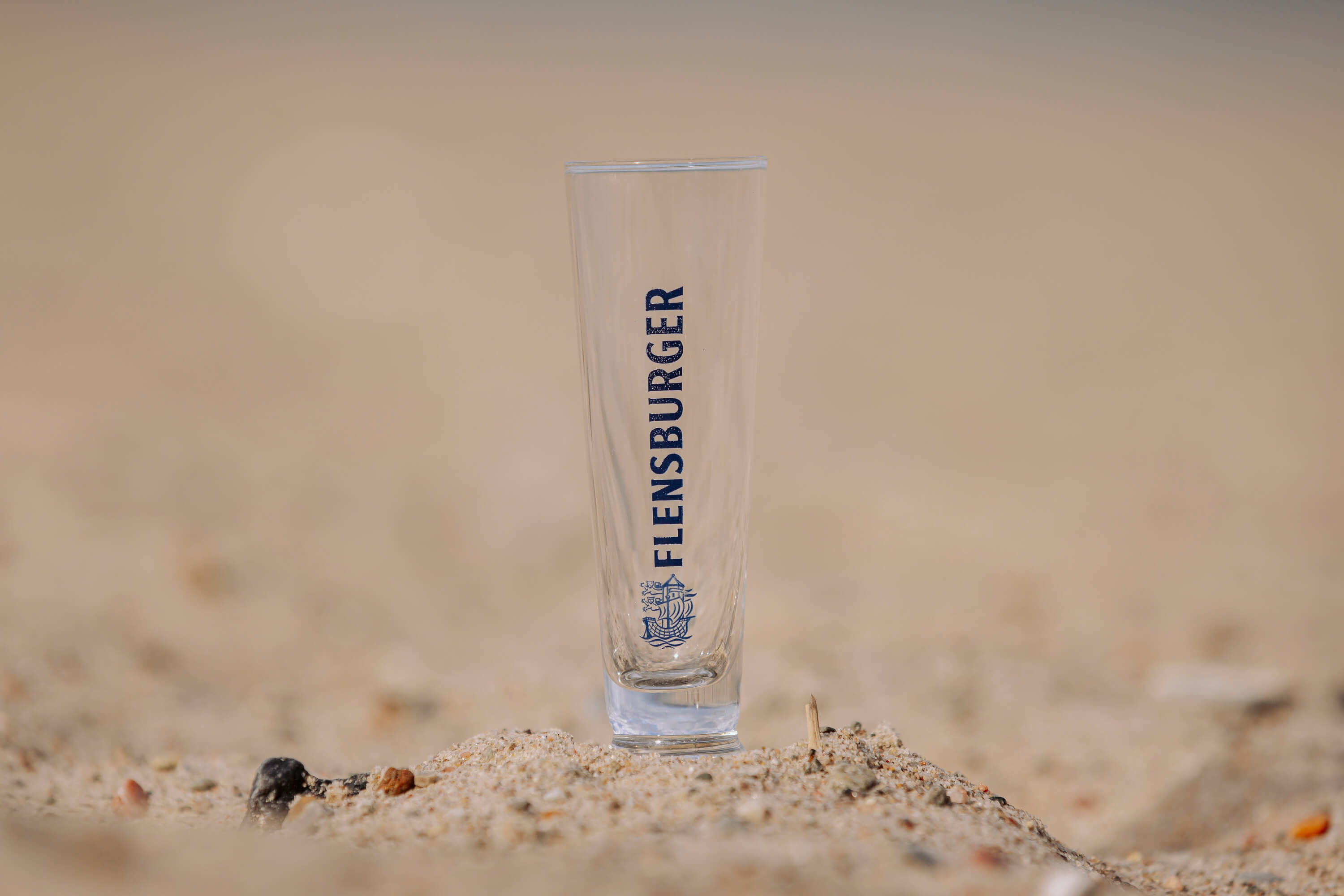 Ein Glas mit der Aufschrift „FLENSBURGER“ steht auf Sand. Das Glas ist schlank und transparent und trägt unten ein blaues Logo mit einem Schiff.