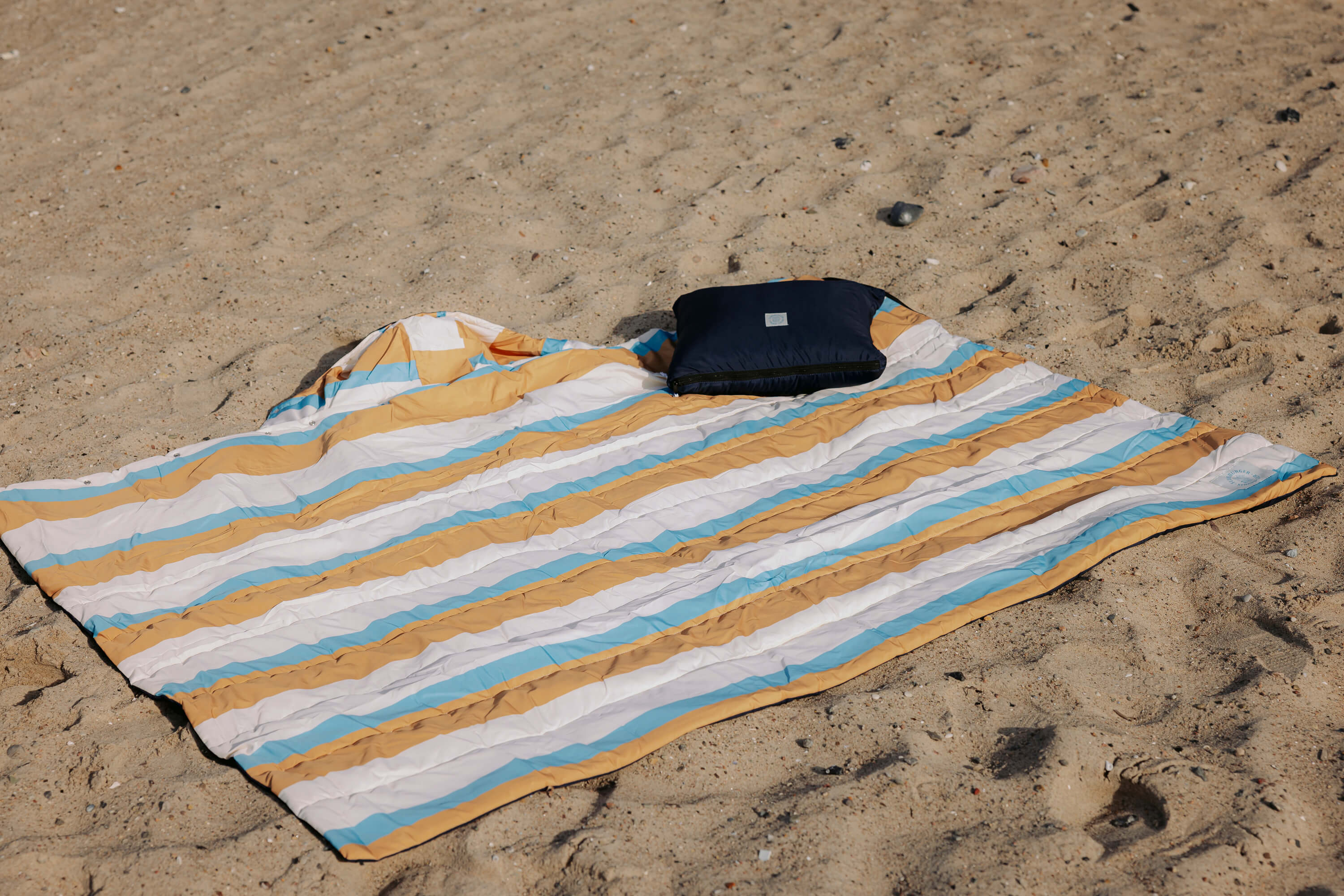 Outdoor-Decke mit Streifen in Beige, Braun, Weiß und Petrolblau liegt auf Sand. Darauf liegt ein zusammengefaltetes, dunkelblaues Kissen.