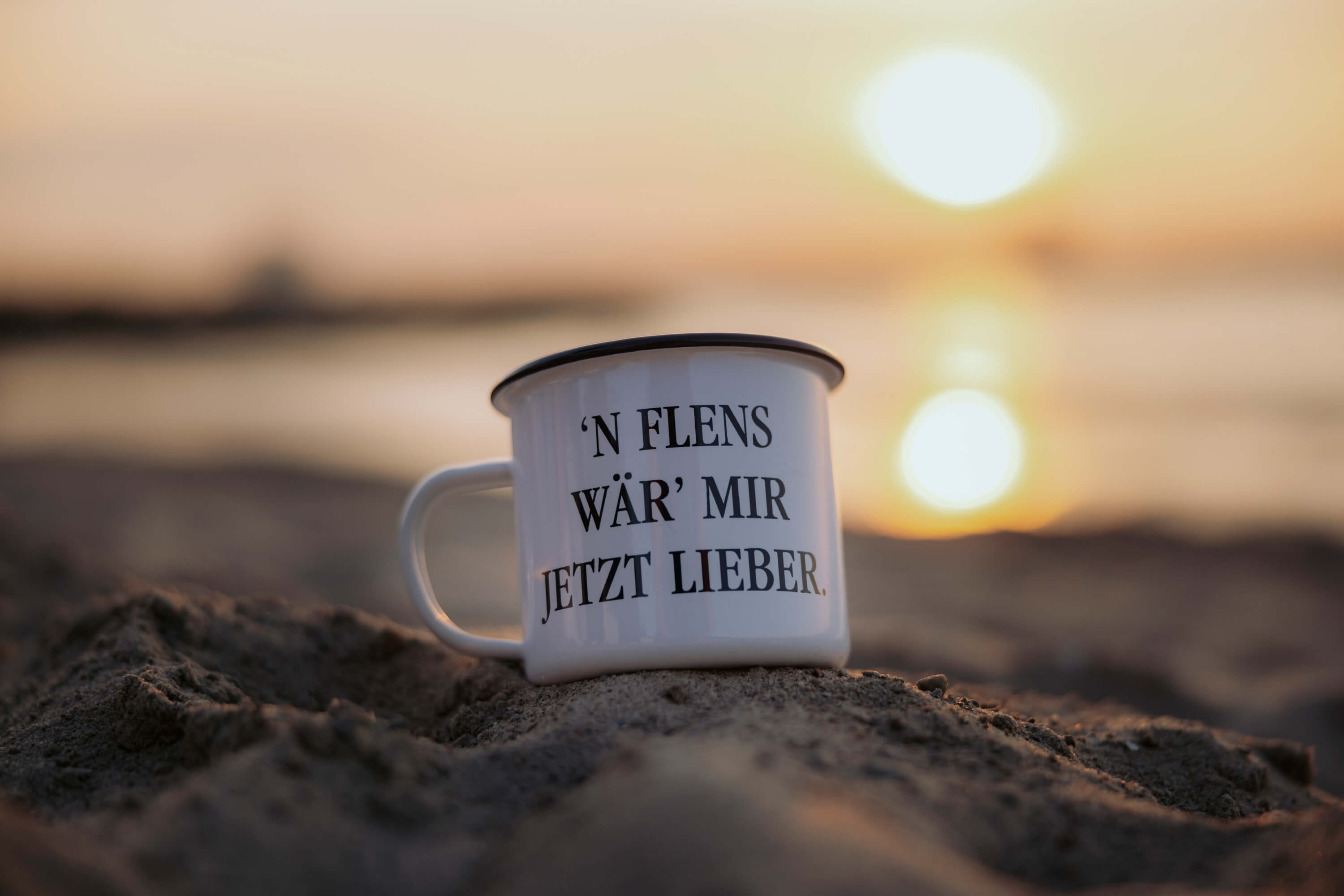 Weiße Emailletasse mit schwarzem Rand steht im Sand vor Sonnenuntergang. Auf der Tasse steht der Text: "'N FLENS WÄR' MIR JETZT LIEBER."