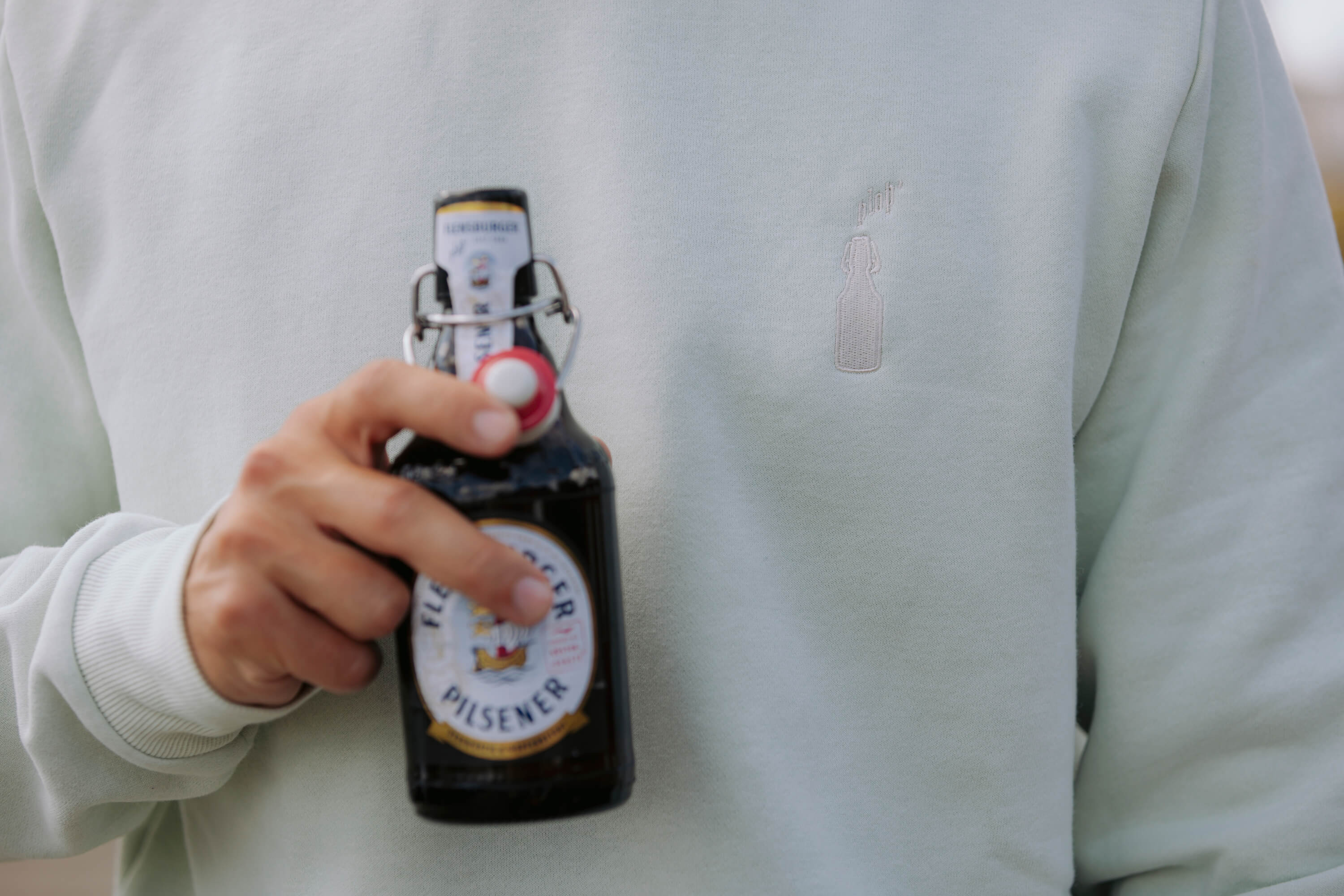 Person hält eine Bierflasche mit Bügelverschluss der Marke "Flensburger Pilsener". Die Person trägt einen mintgrünen Pullover mit einer bestickten Abbildung einer Bierflasche.