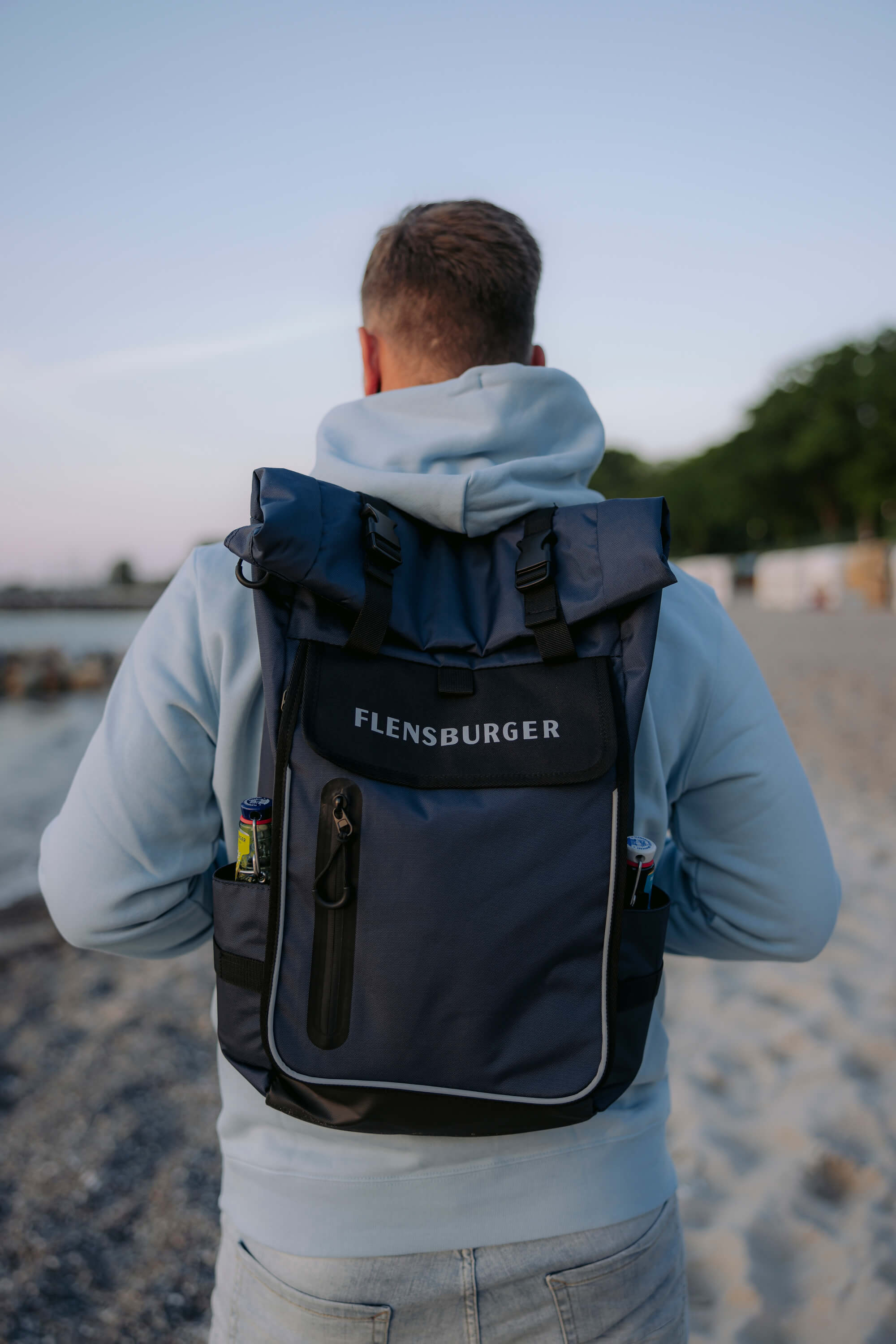 Ein Mann trägt am Strand einen dunkelblauen Rucksack mit dem weißen Schriftzug „FLENSBURGER“ auf dem Rücken. Der Rucksack hat zwei seitliche Taschen mit Flaschen darin, einen Rollverschluss und ein Reißverschlussfach an der Vorderseite.