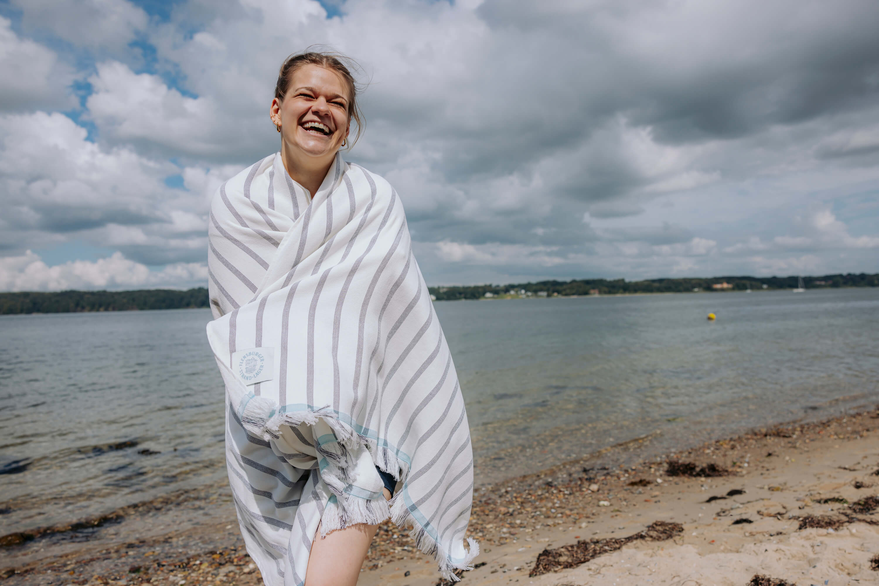Eine Frau steht am Strand und lacht. Sie ist in ein weißes Handtuch mit grauen Streifen und Fransen am unteren Ende gehüllt.