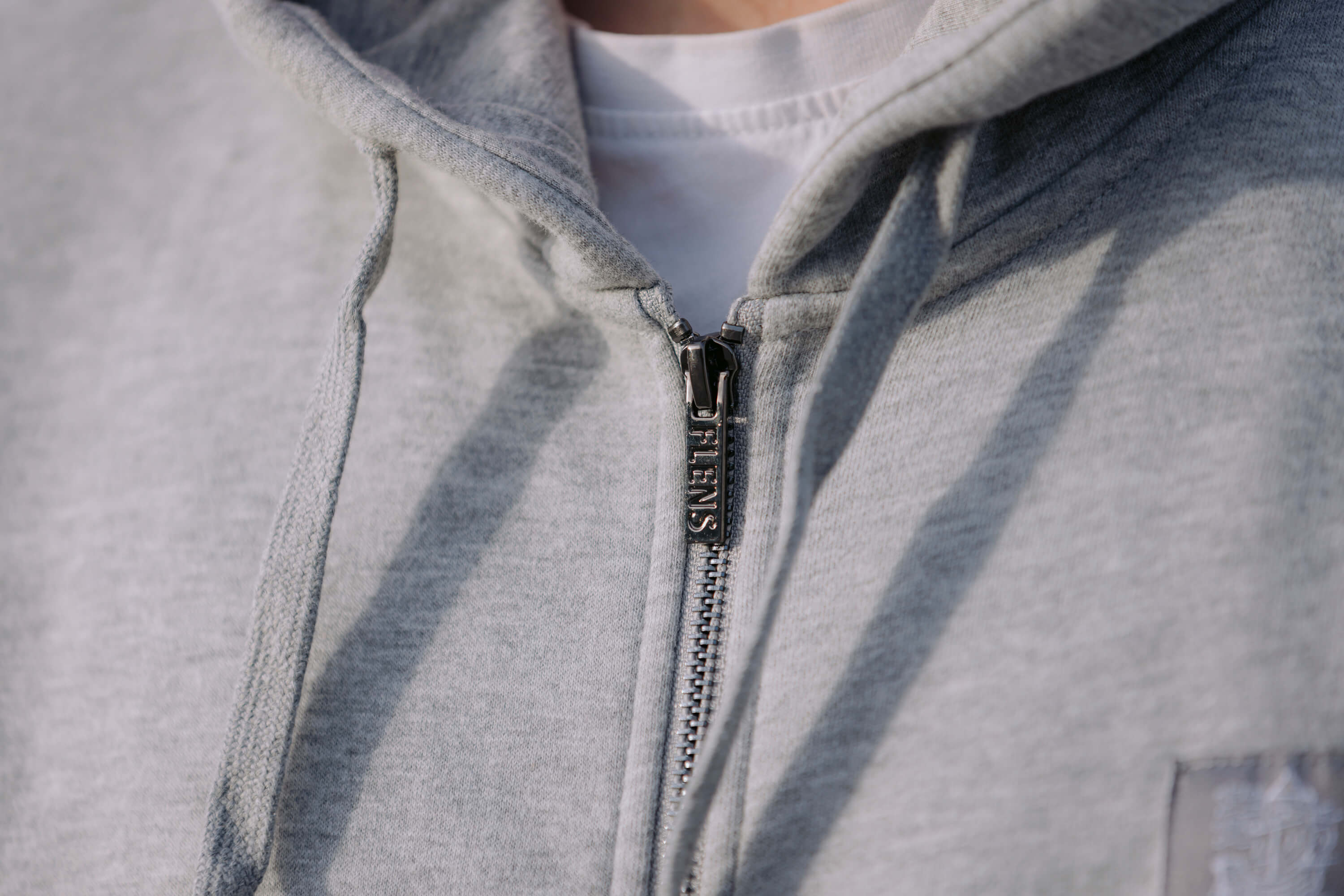 Nahaufnahme eines Reißverschlusses an einer hellgrauen Sweatshirtjacke mit der Aufschrift „FLENS“ auf dem Zipper. Die Jacke hat eine Kapuze und zwei Kordeln.