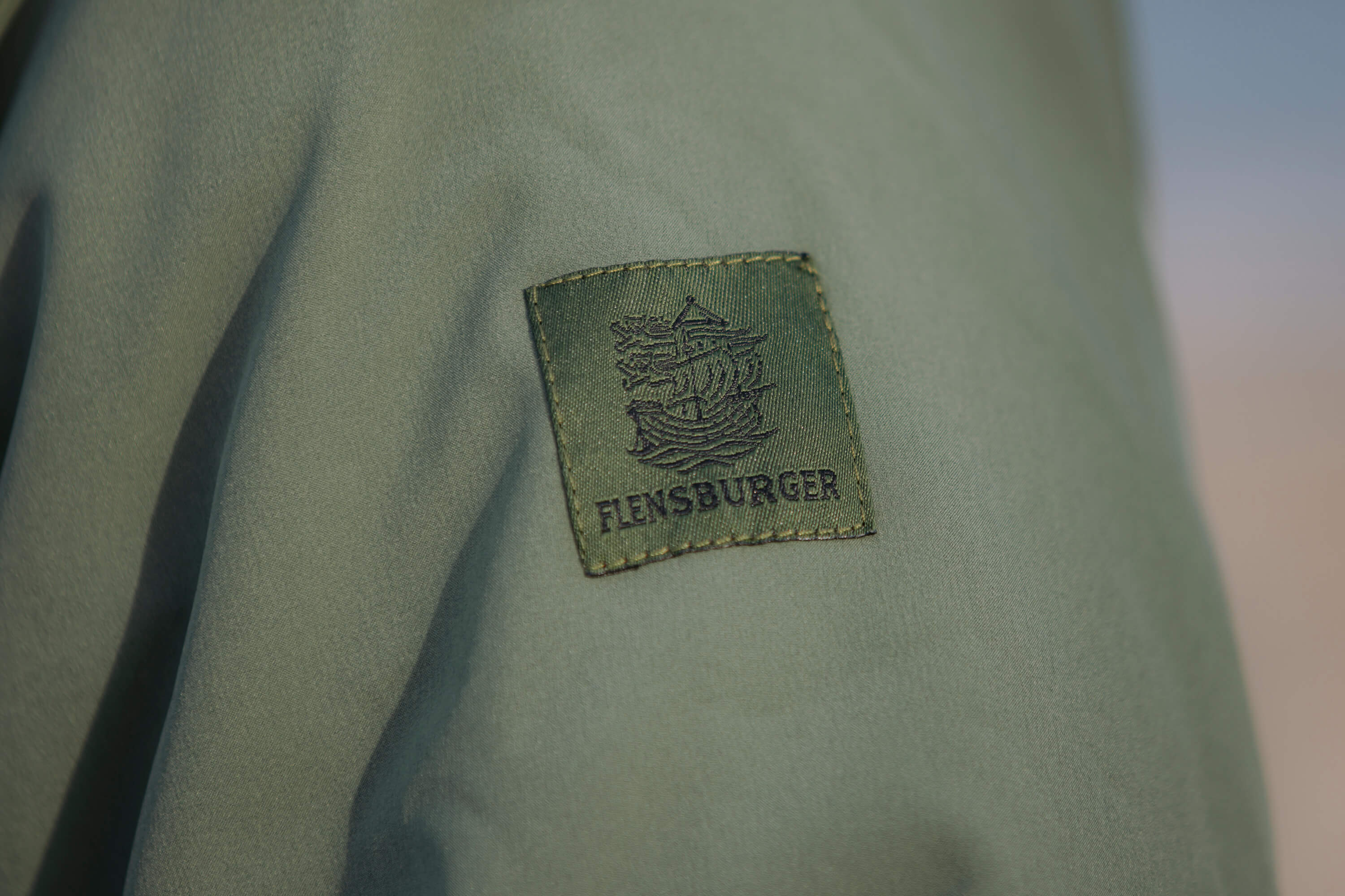 Detailansicht eines grünen Windbreakers zeigt ein rechteckiges, gesticktes Logo mit einem Segelschiff und der Schrift „FLENSBURGER“ auf dem Ärmel. Das Material wirkt wetterfest und glatt.