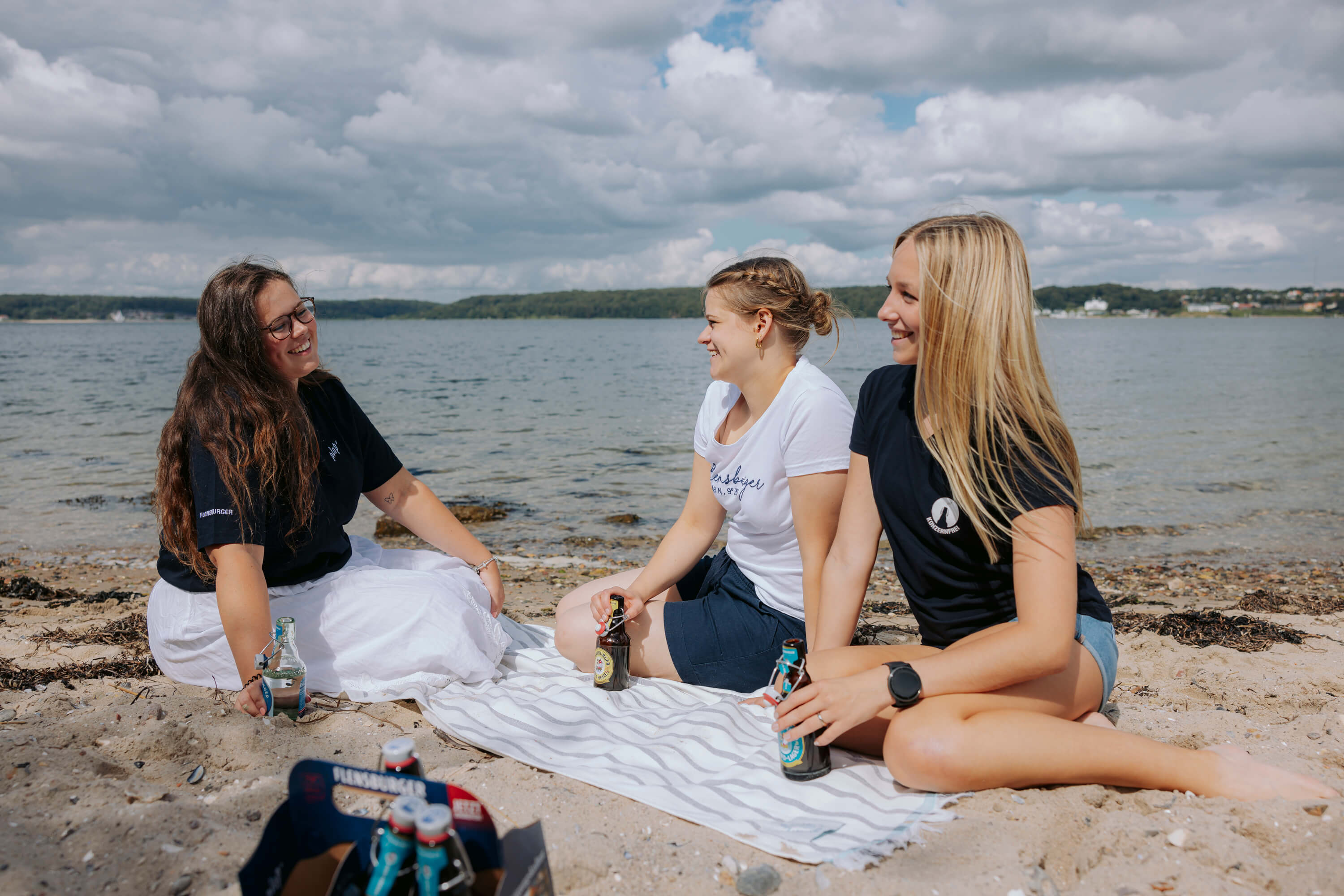 Drei junge Frauen sitzen auf einer weiß-grau gestreiften Decke am Strand. Sie lachen und halten Flaschen mit Getränken in den Händen, während im Hintergrund das Meer und ein bewölkter Himmel zu sehen sind.