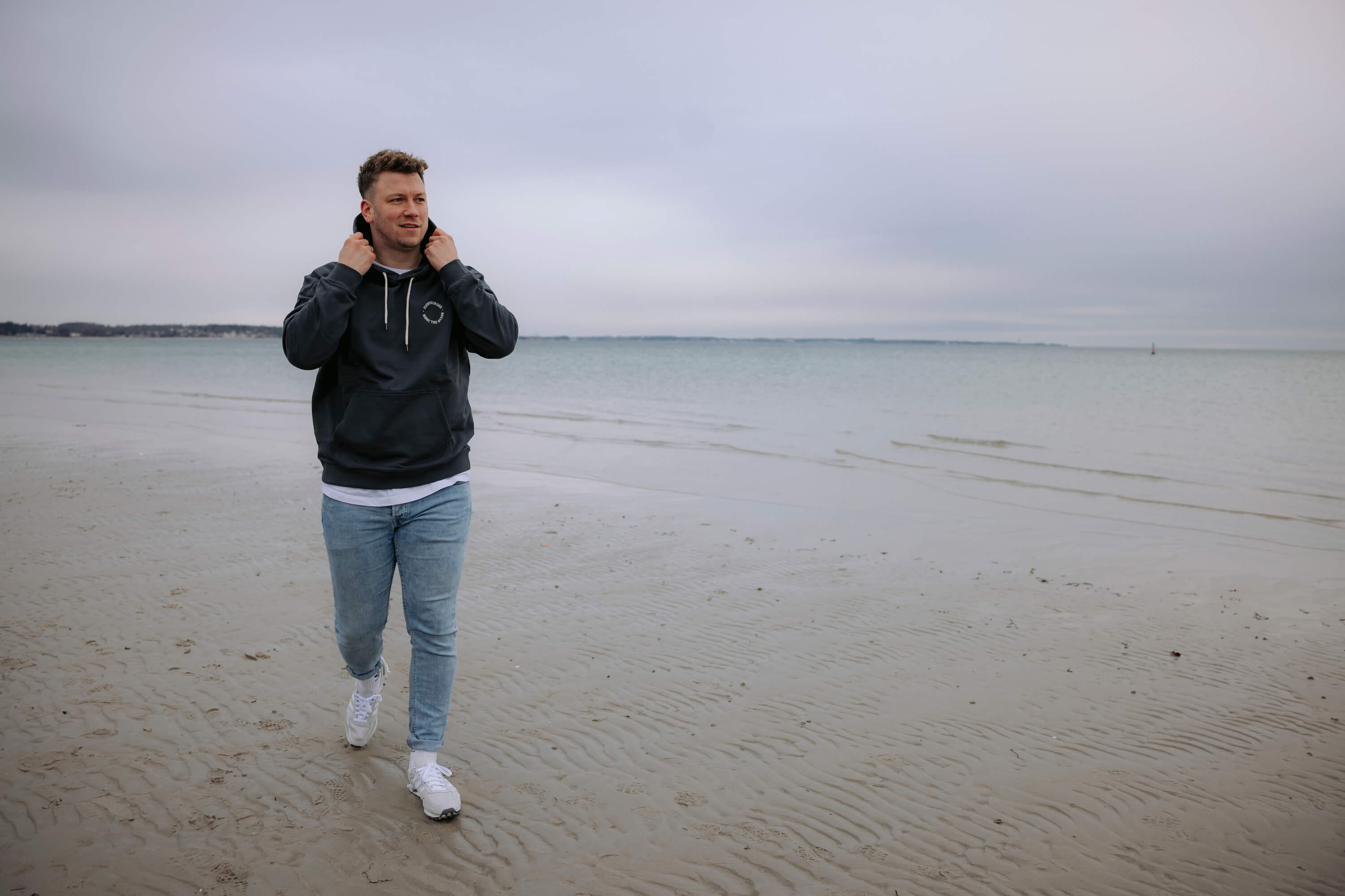 Ein Mann trägt den anthrazitfarbenen Hoodie "Embrace the Ocean". Er geht am Strand und hält beide Hände an seine Kapuze.