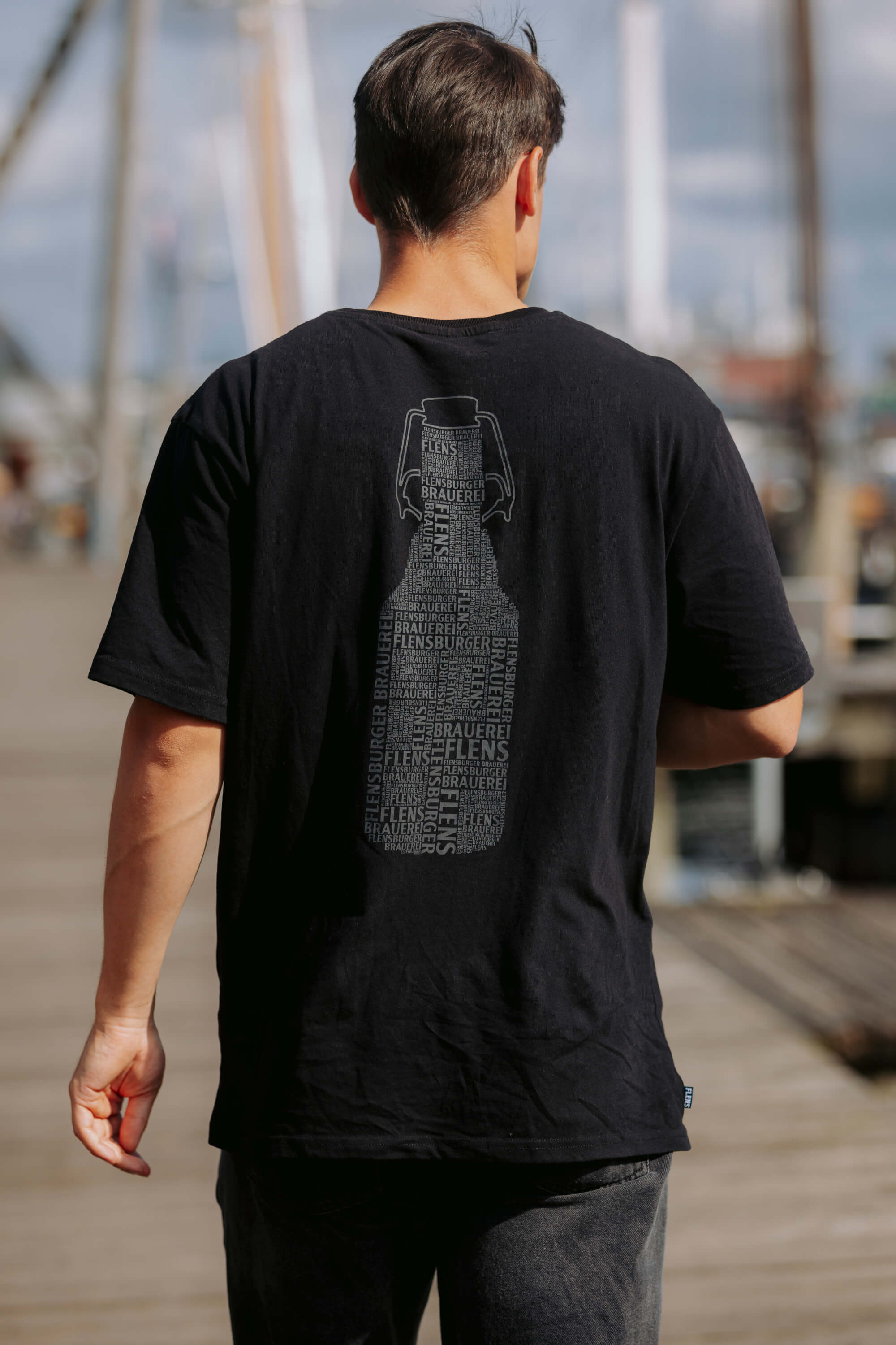 Mann trägt ein schwarzes T-Shirt mit einem großen Backprint einer Bügelverschlussflasche aus Wörtern wie „FLENS“ und „BRAUEREI“ in verschiedenen Größen und Ausrichtungen. Im Hintergrund ist ein Hafen zu sehen.