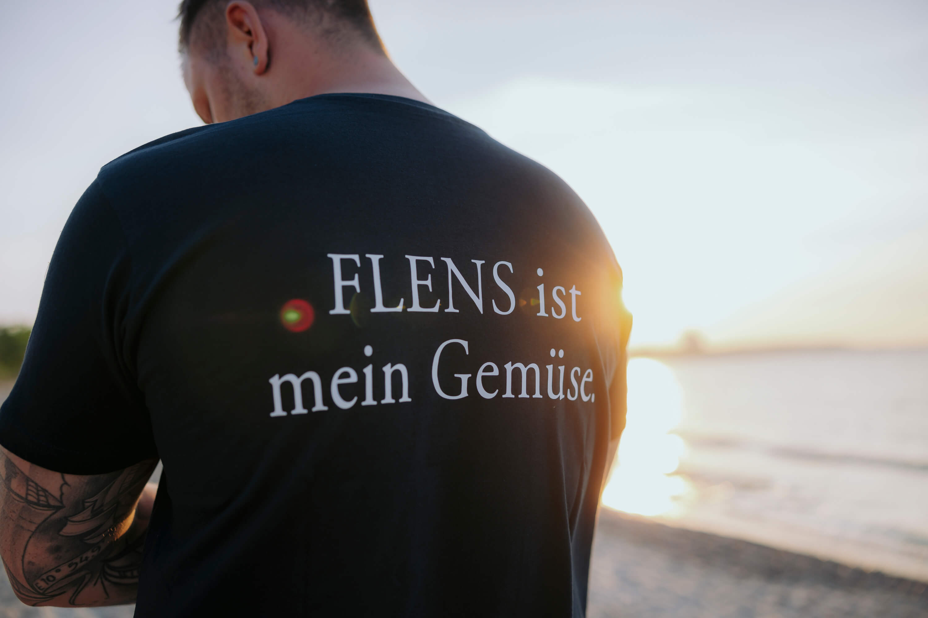 Ein Mann steht am Strand mit Blick auf das Wasser bei Sonnenuntergang. Auf seinem schwarzen T-Shirt steht in weißen Buchstaben: „FLENS ist mein Gemüse.“ auf dem oberen Rücken.