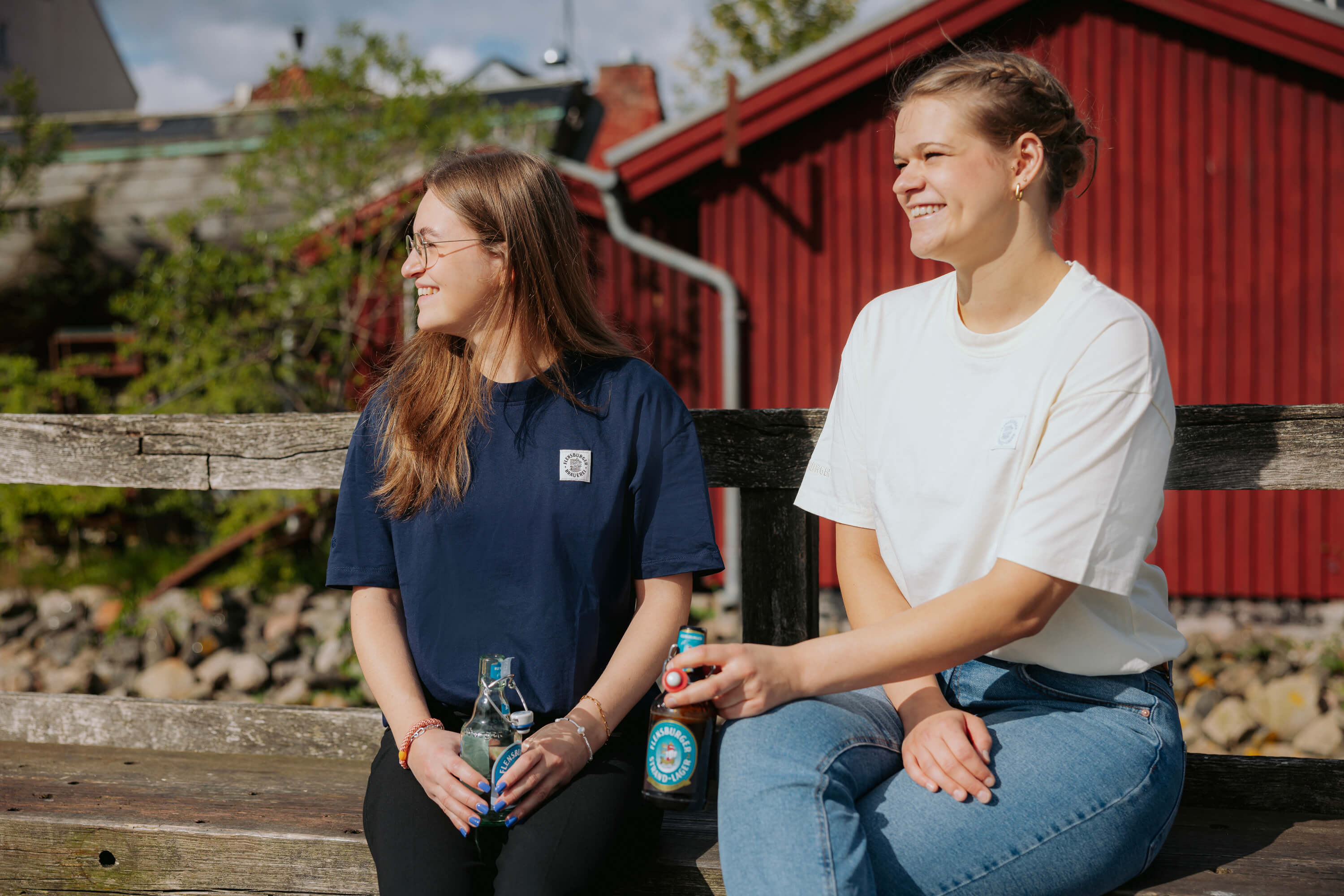 Zwei junge Frauen sitzen auf einer Bank vor einem roten Holzhäuschen. Die linke trägt ein navyblaues T-Shirt, die rechte ein weißes T-Shirt. beide lächeln und halten jeweils eine Flensburger Bügeverschlussflasche.