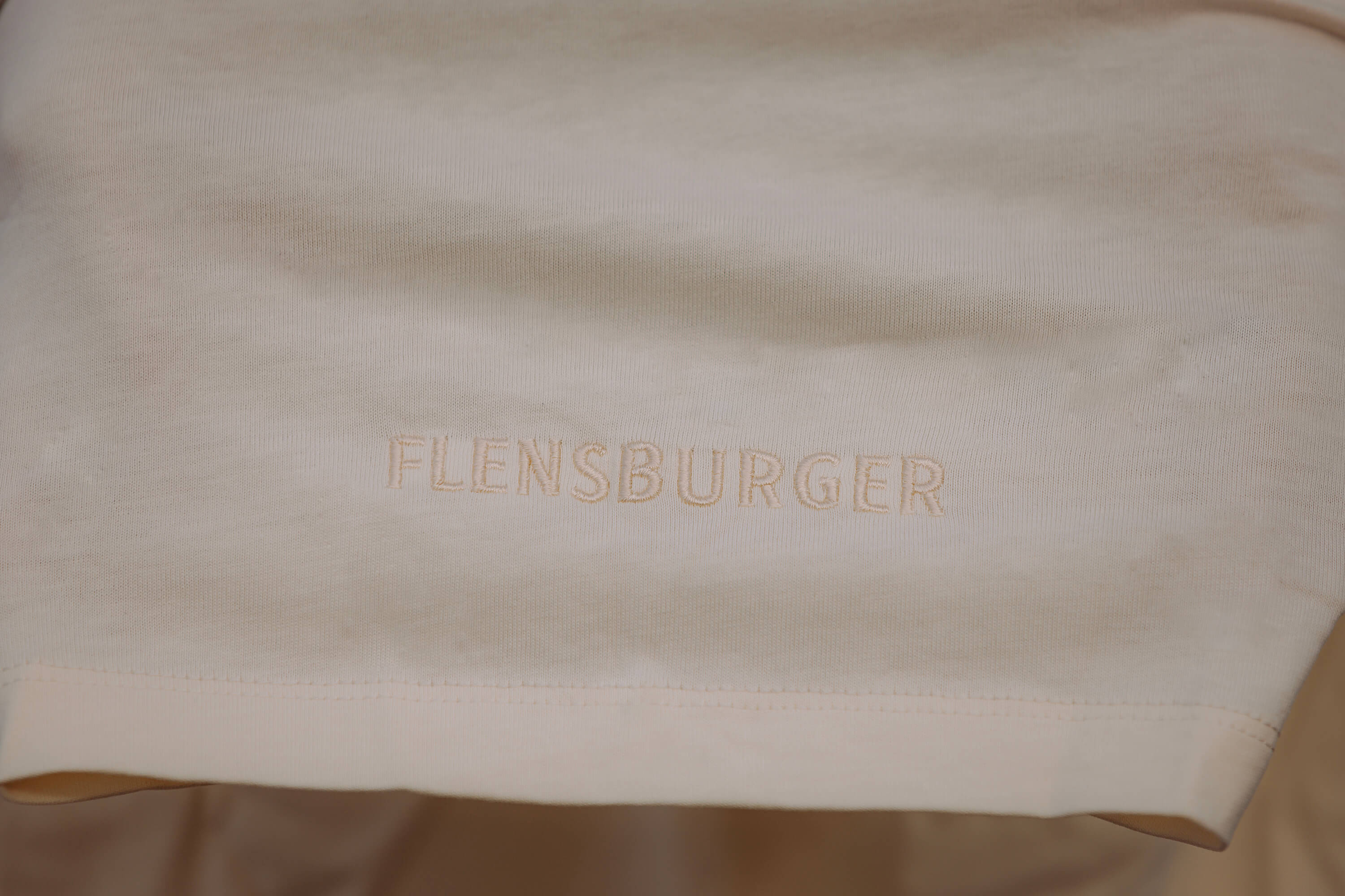 Detailansicht: Beige hellblaues Kurzarmshirt mit gesticktem Schriftzug „FLENSBURGER“ im Vordergrund.