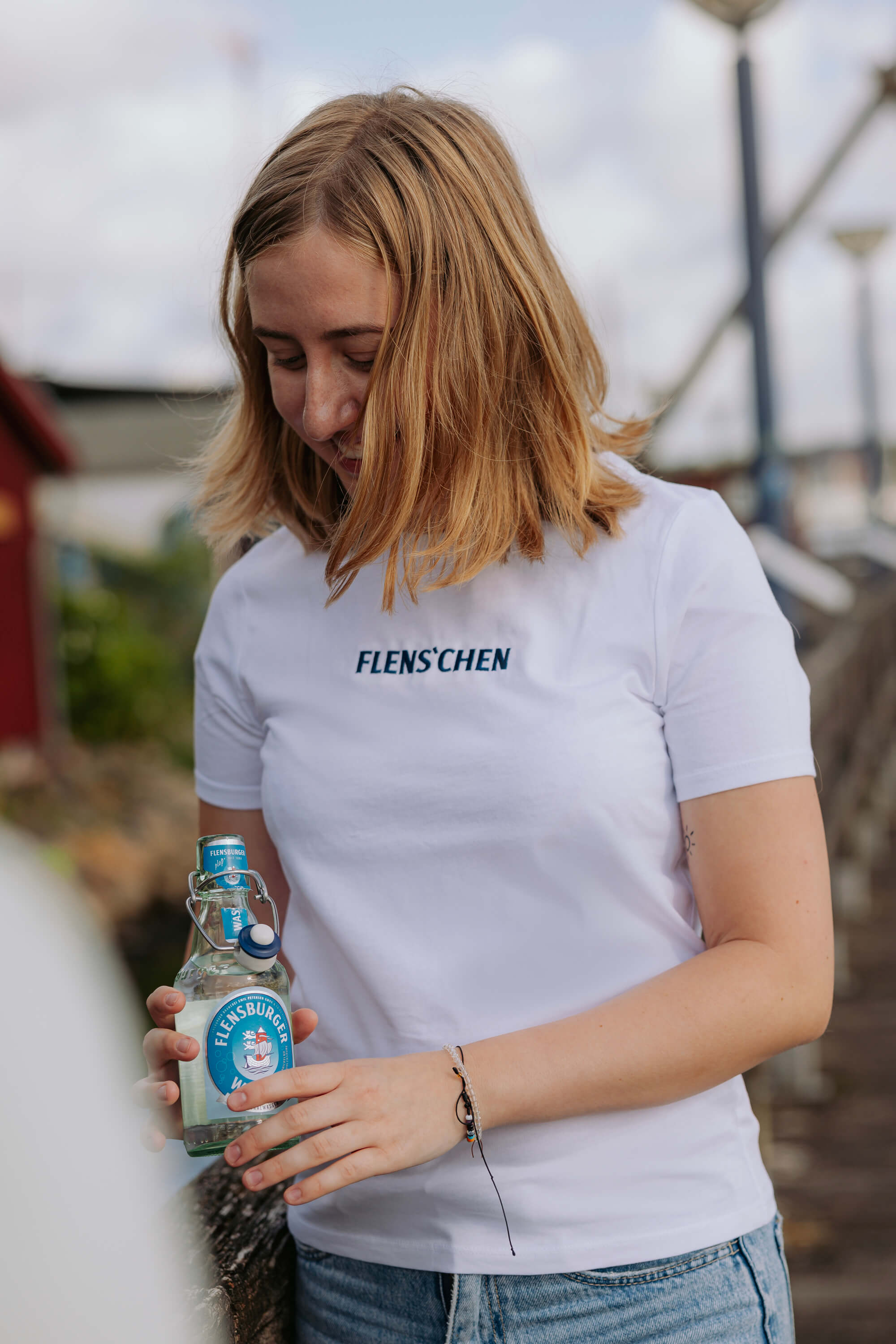 Junges Frau trägt ein weißes T-Shirt mit der Aufschrift "FLENS'CHEN". Sie hält eine klare Flensburger-Wasser-Flasche mit blauem Etikett. Sie steht draußen.