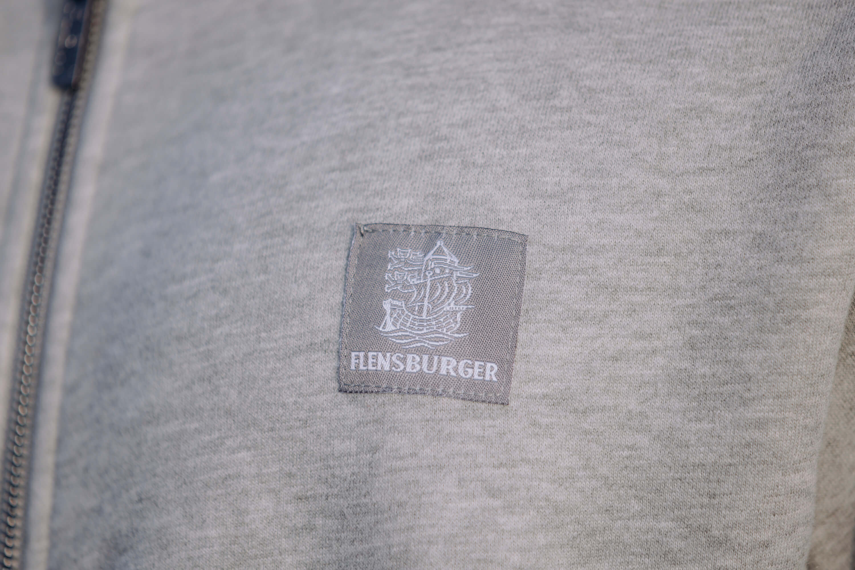 Ein graues Sweatshirt zeigt ein kleines, graues Stoffetikett mit weißem Segelschiff und dem Schriftzug "FLENSBURGER". Das Material wirkt weich und ist leicht meliert.