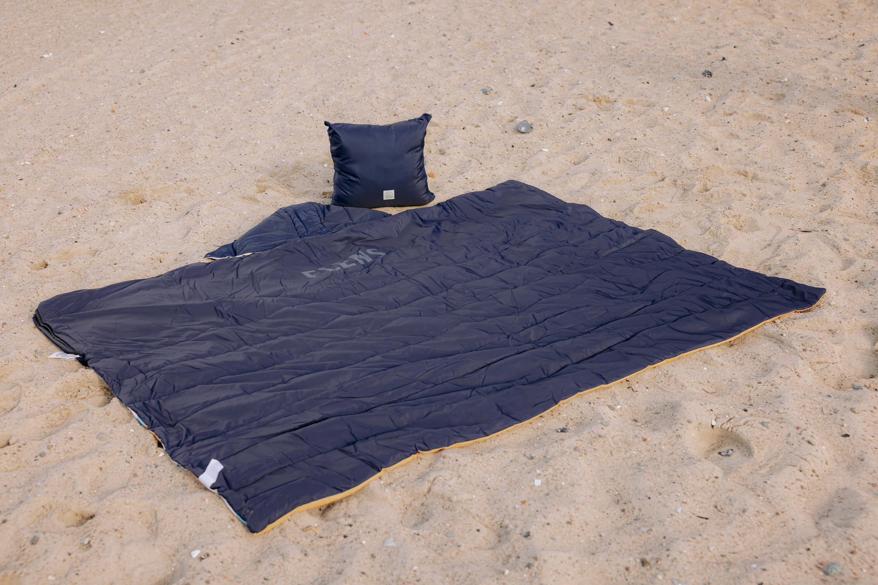 Dunkelblaue Outdoordecke liegt offen im Sand. Ein passendes, ebenfalls dunkelblaues Kissen steht aufgerichtet am oberen Rand der Decke.