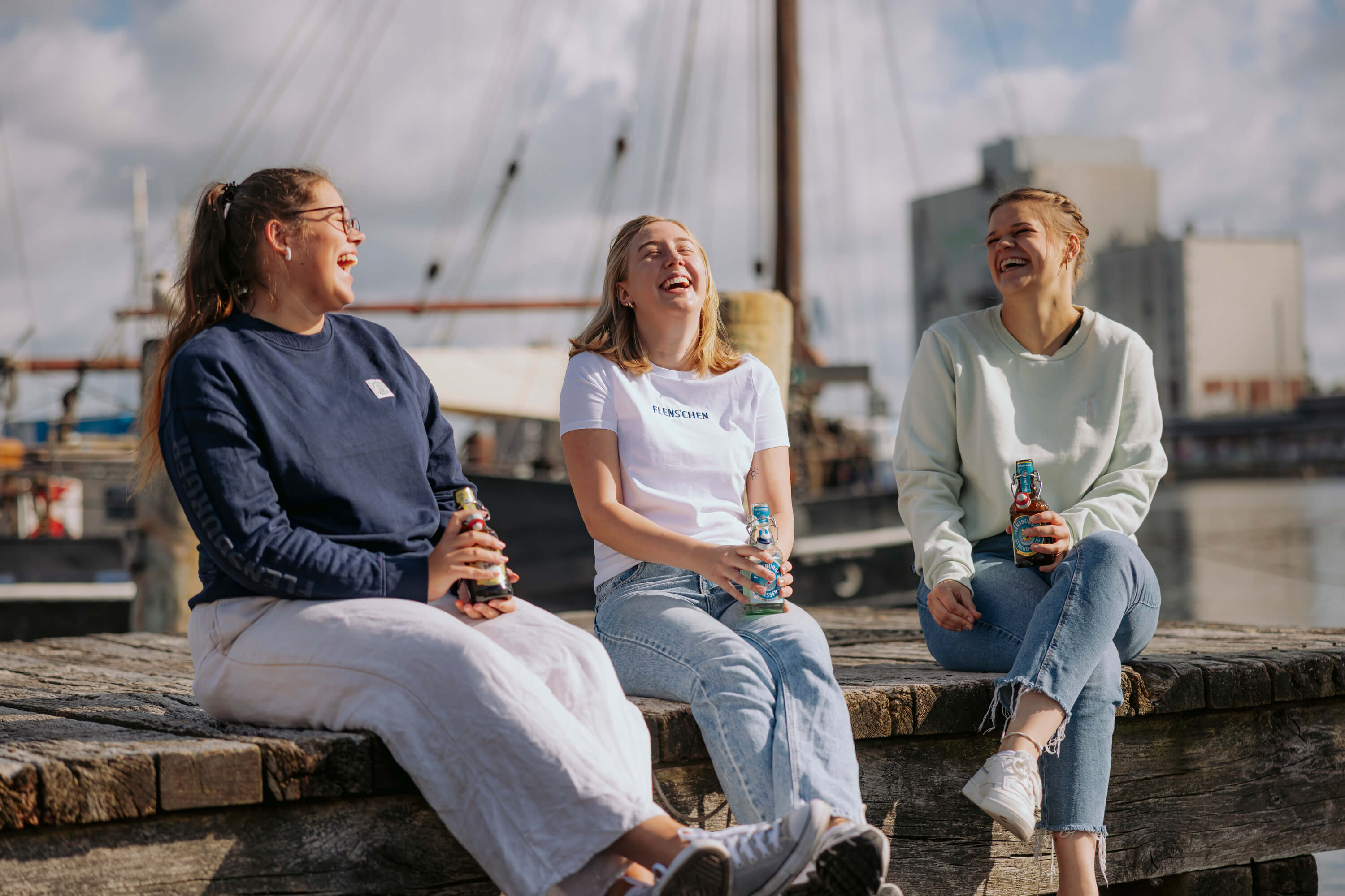 Drei junge Frauen sitzen lachend auf einer Holzsteg am Wasser. Jede hält eine Flasche in der Hand, im Hintergrund sind Boote und ein Hafen zu sehen.