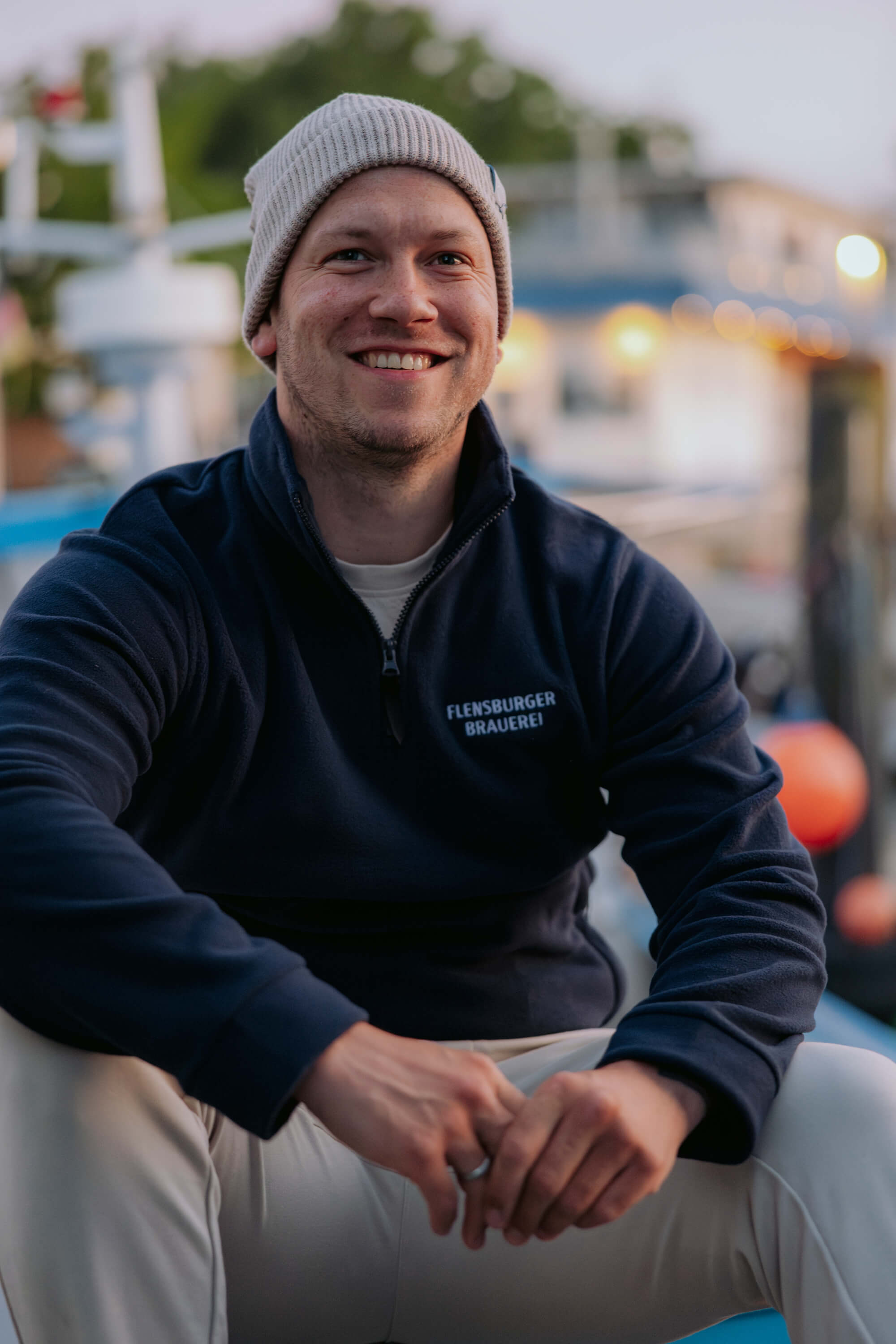 Ein Mann trägt einen navyblauen Fleece-Pullover mit der Aufschrift „FLENSBURGER BRAUEREI“ und eine beige Strickmütze. Er sitzt entspannt im Freien bei Dämmerung.
