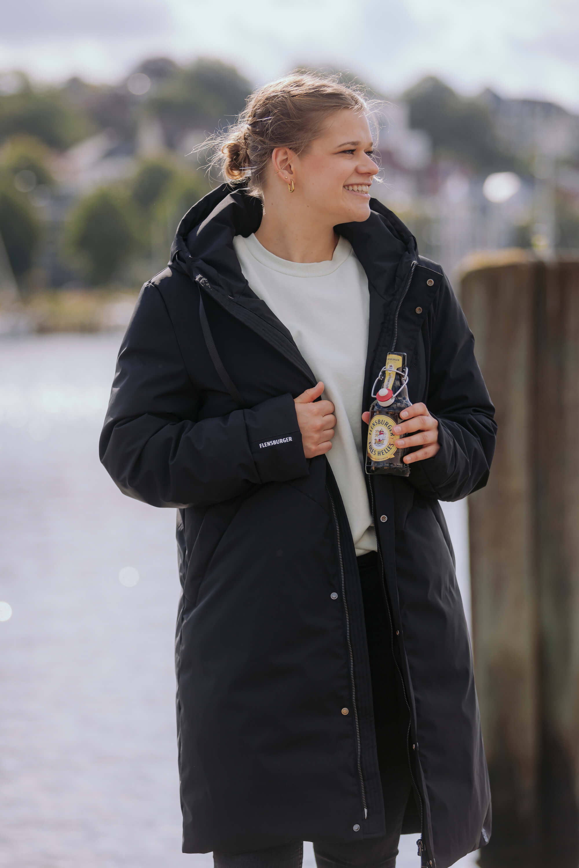 Eine Frau trägt den schwarzen FLENS Winterparka "Ruben" und hält eine Flasche Flensburger Pils Helles. Sie steht lächelnd vor einem Gewässer.
