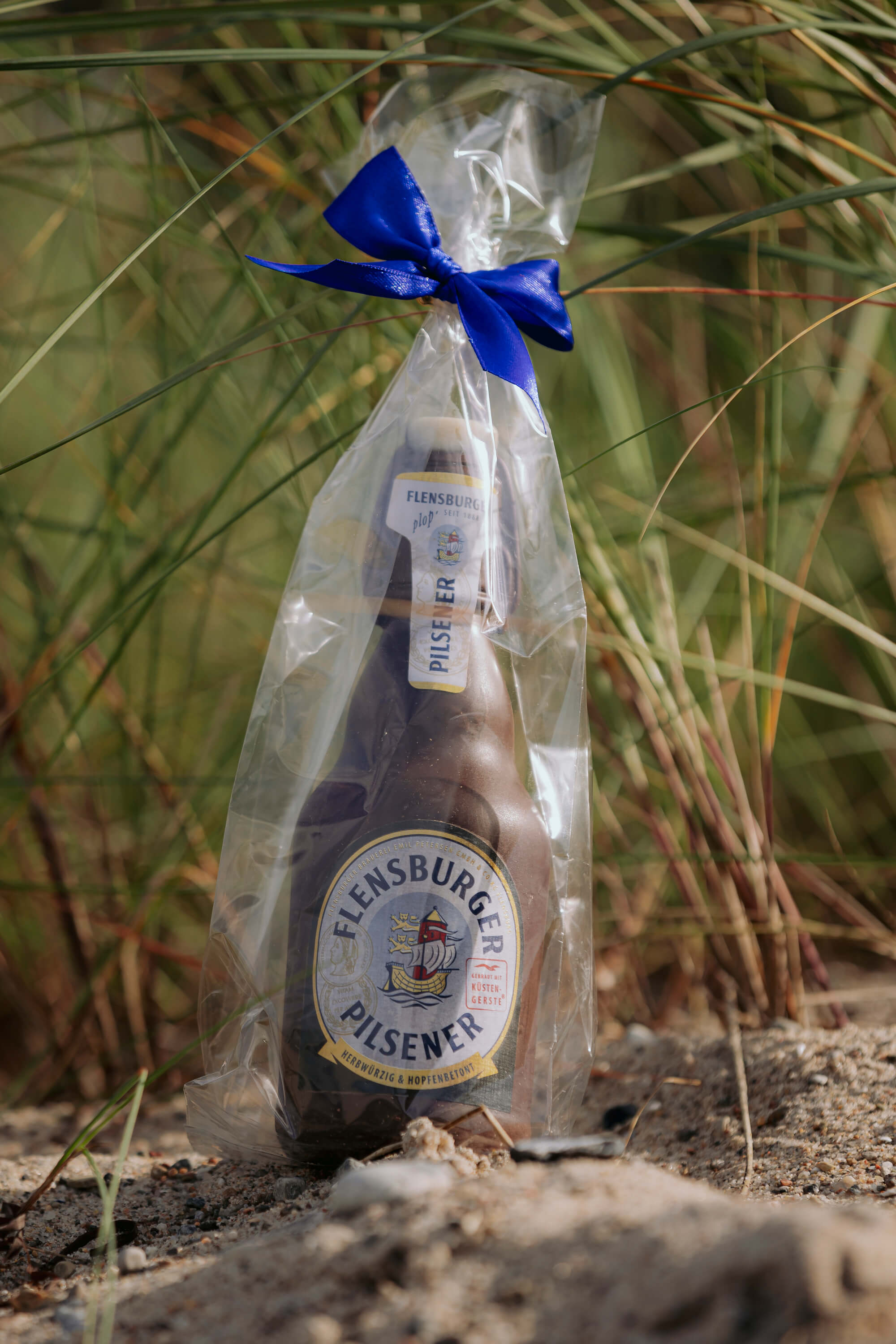 Flensburger Pilsener Schokoladen-Flasche in 0,3 l, verpackt in durchsichtigem Plastik mit blauem Schleifenband, steht auf sandigem Boden vor grünen Gräsern.