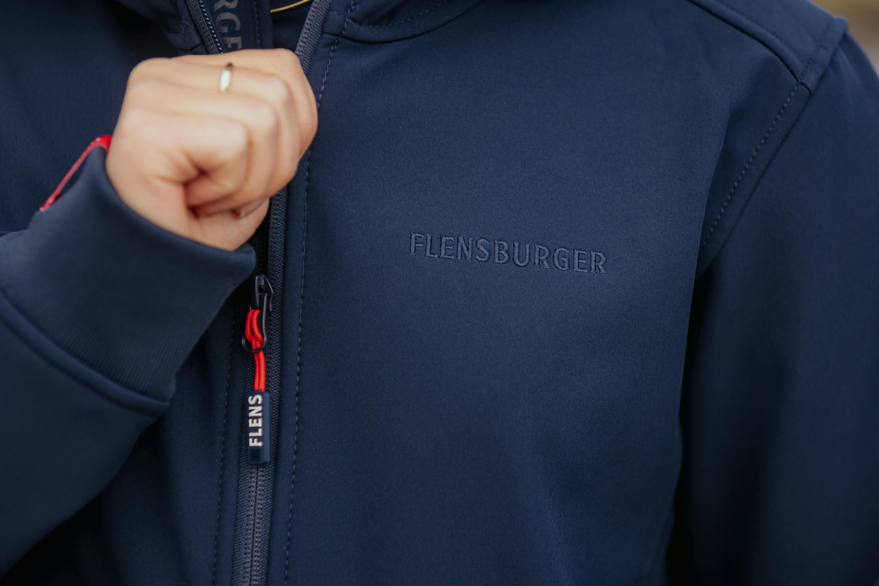 Eine Frau hält den Reißverschluss der navyblauen Softshelljacke. Auf der Brust steht dezent „FLENSBURGER“. Am Reißverschlussanhänger steht „FLENS“.