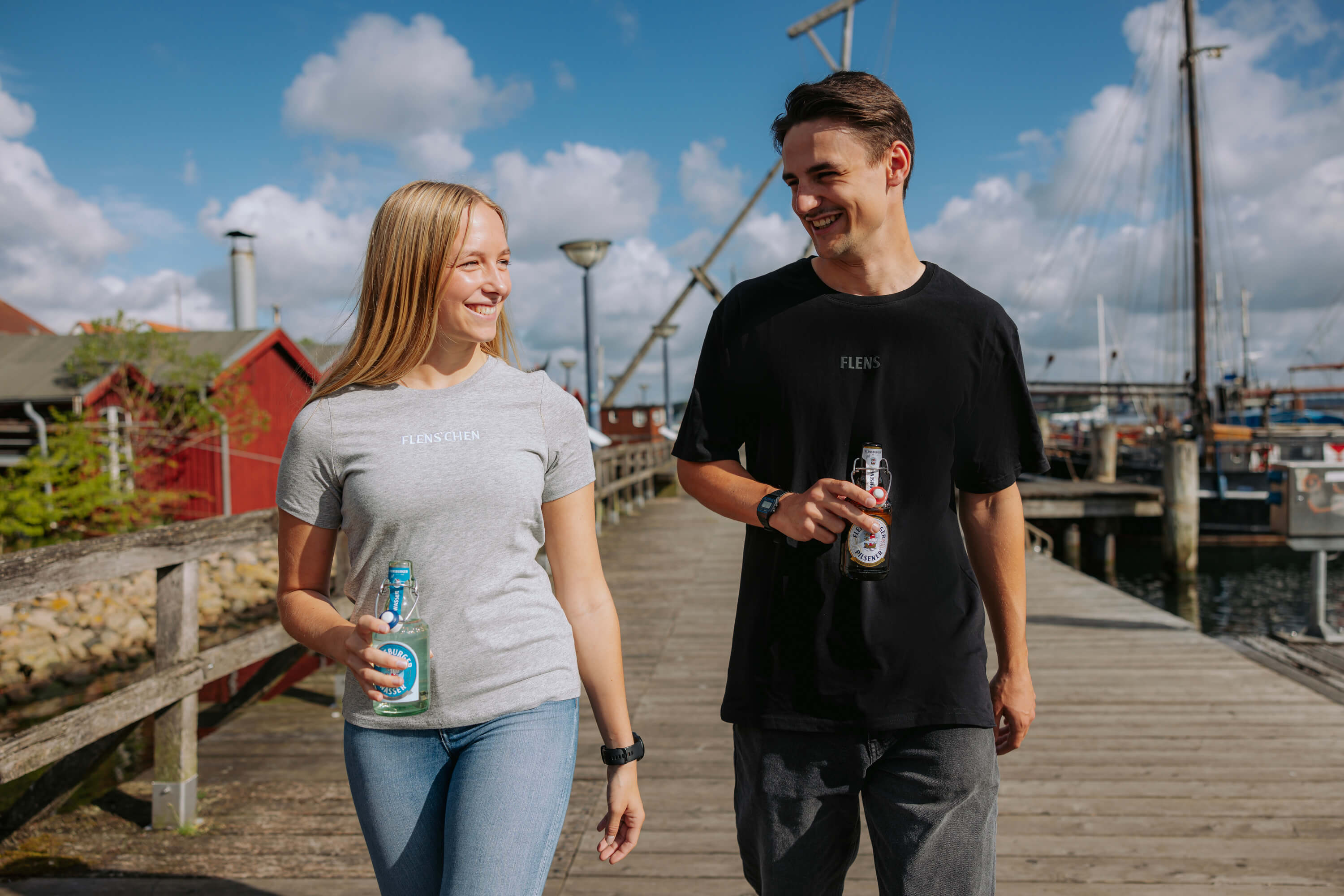 Eine Frau trägt ein graues T-Shirt mit der Aufschrift „FLENS’CHEN“ und hält eine Flasche. Ein Mann trägt ein schwarzes T-Shirt mit der Aufschrift „FLENS“ und hält ebenfalls eine Flasche. Beide gehen auf einem Steg am Hafen.