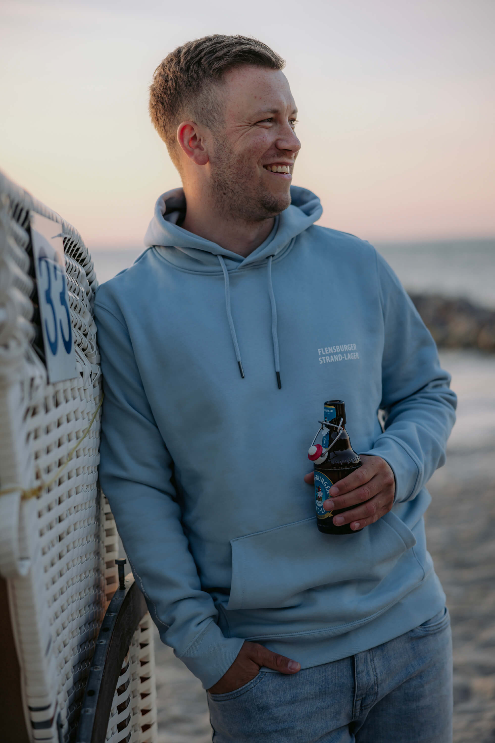 Ein Mann lehnt an einem weißen Strandkorb. Er trägt einen hellblauen Hoodie mit dem Schriftzug „Flensburger Strand-Lager“ und hält eine Flasche Strand-Lager-Bier in der Hand.