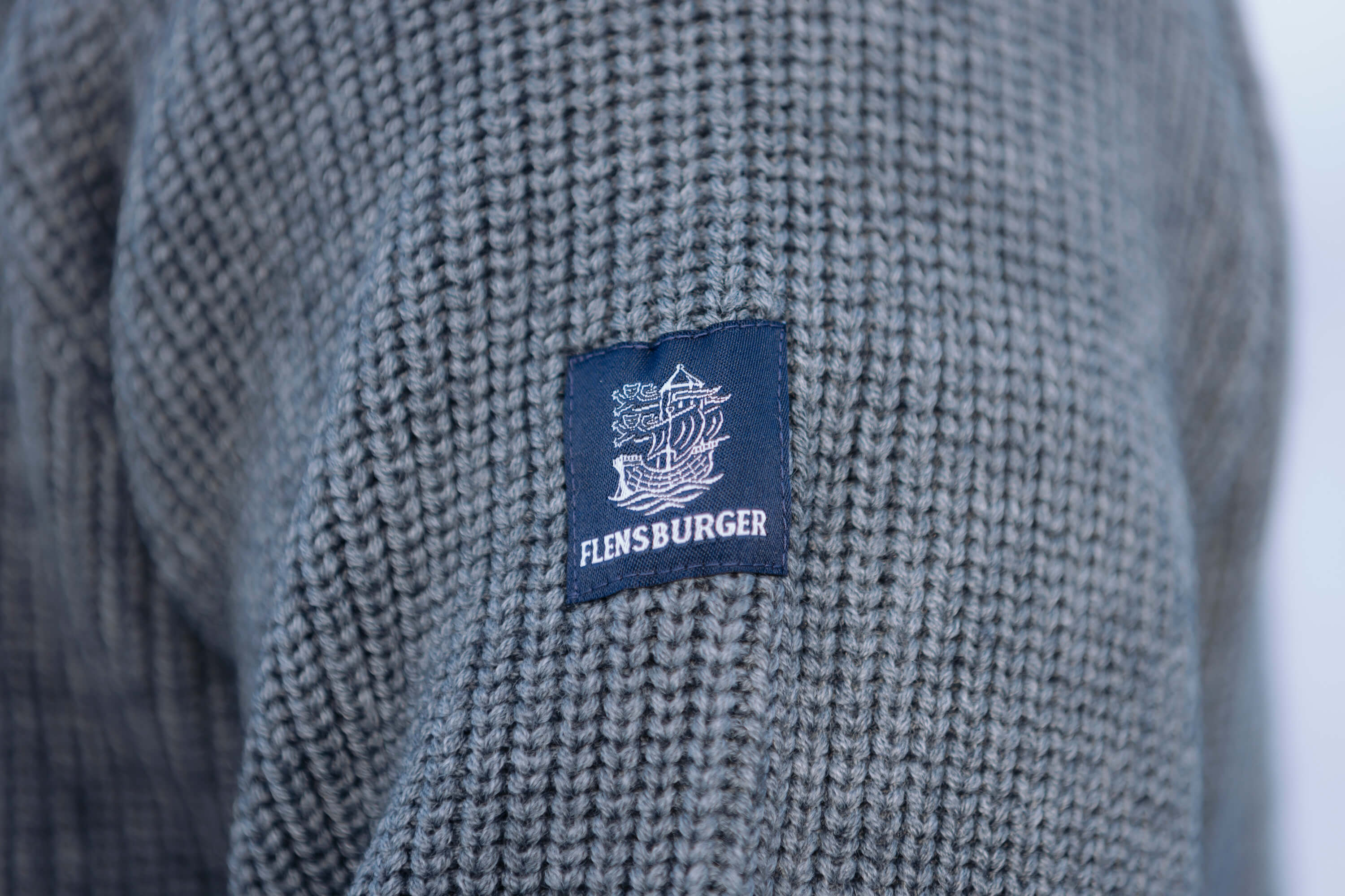 Blaues Stofflabel mit weißem Segelschiff-Logo und dem Schriftzug „FLENSBURGER“ auf grauem, grob gestricktem Pullover in Anthrazit.