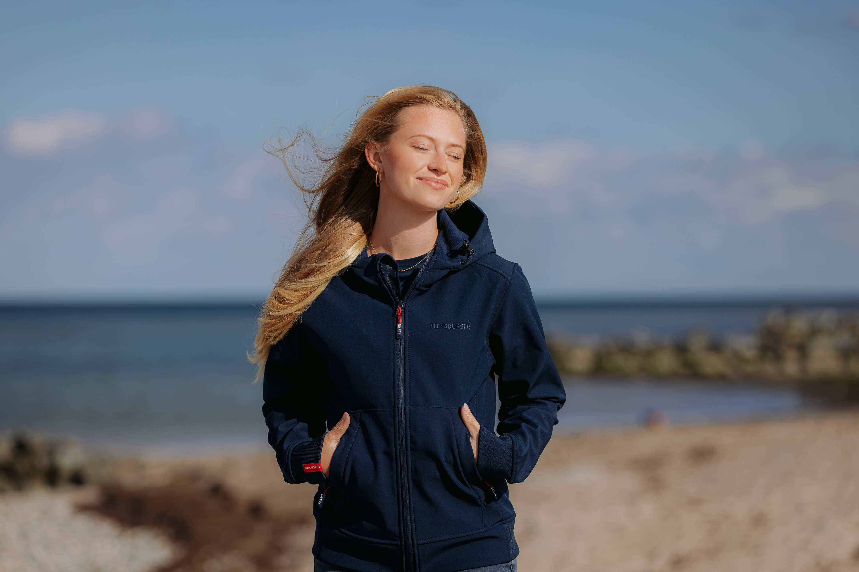 Eine blonde Frau trägt eine navyblaue Softshelljacke mit Kapuze und hält ihre Hände in den Taschen. Sie steht entspannt am Strand mit leicht geschlossenen Augen.