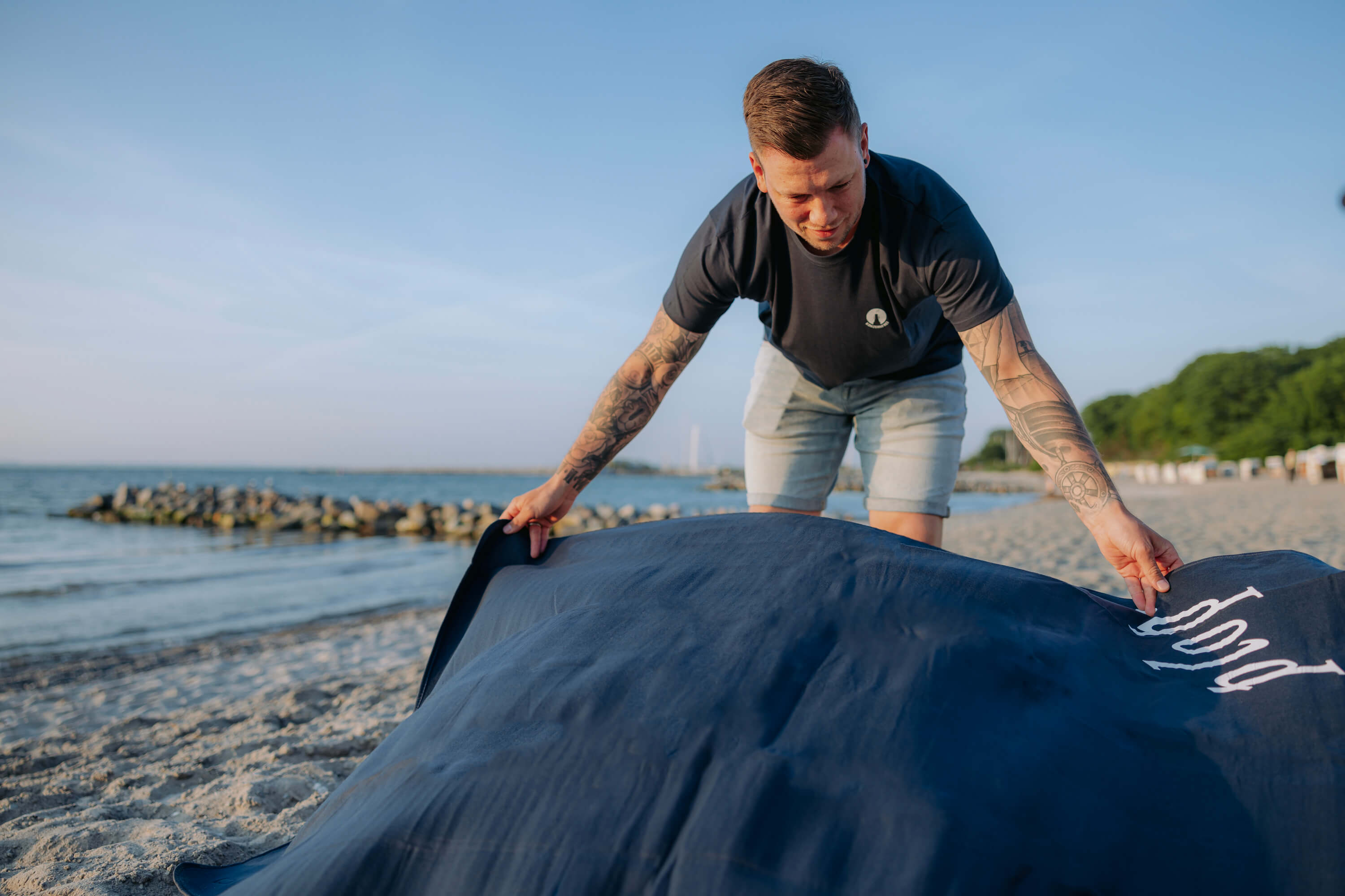 Mann mit tätowierten Armen breitet eine große, dunkelblaue Stranddecke mit dem weißen Schriftzug "plop'" am Sandstrand aus. Im Hintergrund sind Meer, Steine und Bäume sichtbar.