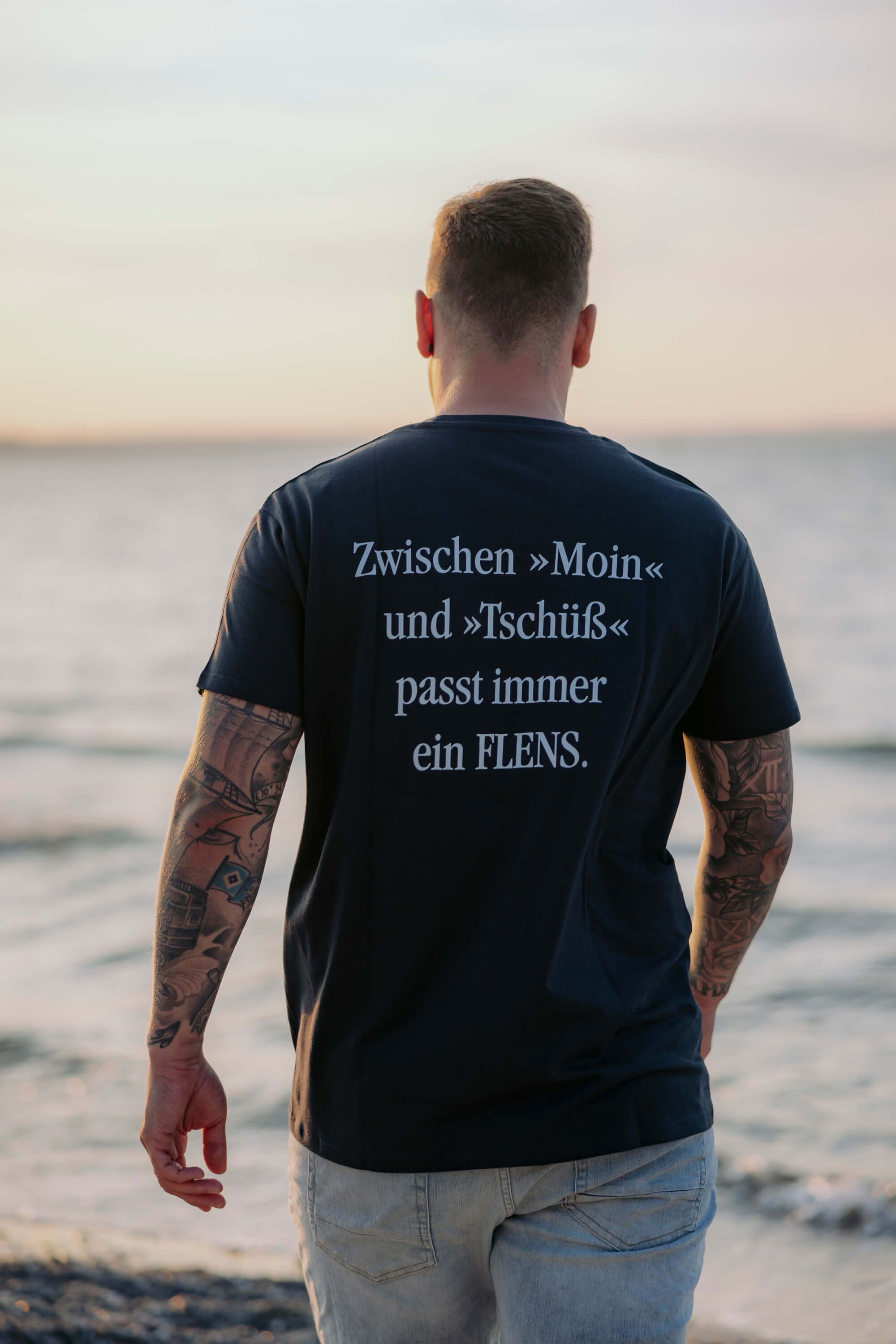 Mann mit Tattoos auf den Armen trägt ein schwarzes T-Shirt mit der Aufschrift: „Zwischen »Moin« und »Tschüss« passt immer ein FLENS.“ Er steht mit dem Rücken zur Kamera am Strand mit Blick aufs Wasser.