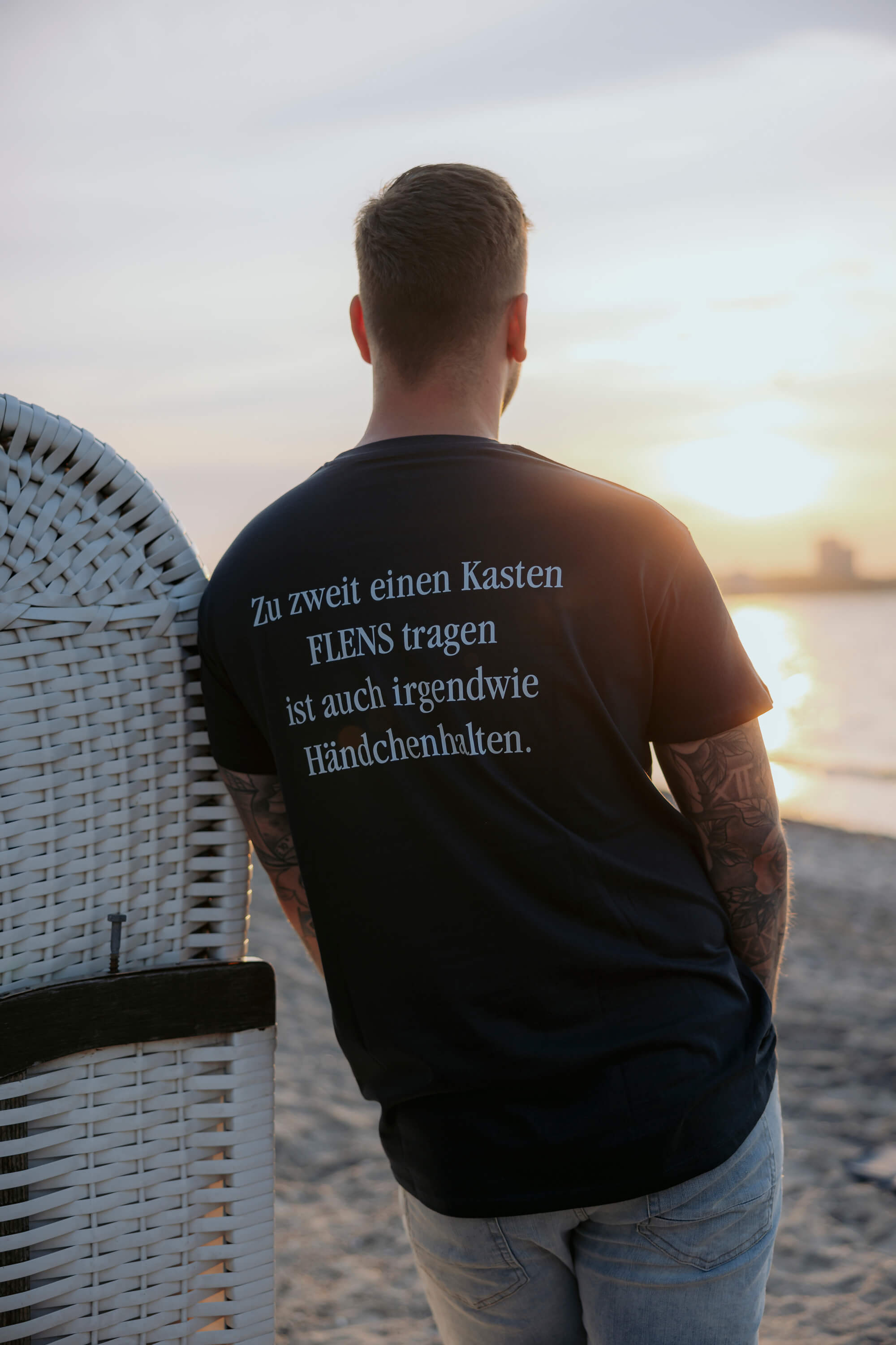 Mann lehnt an einem weißen Strandkorb und trägt ein schwarzes T-Shirt mit dem Text: „Zu zweit einen Kasten FLENS tragen ist auch irgendwie Händchenhalten.“ Er steht mit dem Rücken zur Kamera und im Hintergrund ist ein Sonnenuntergang am Strand zu sehen.