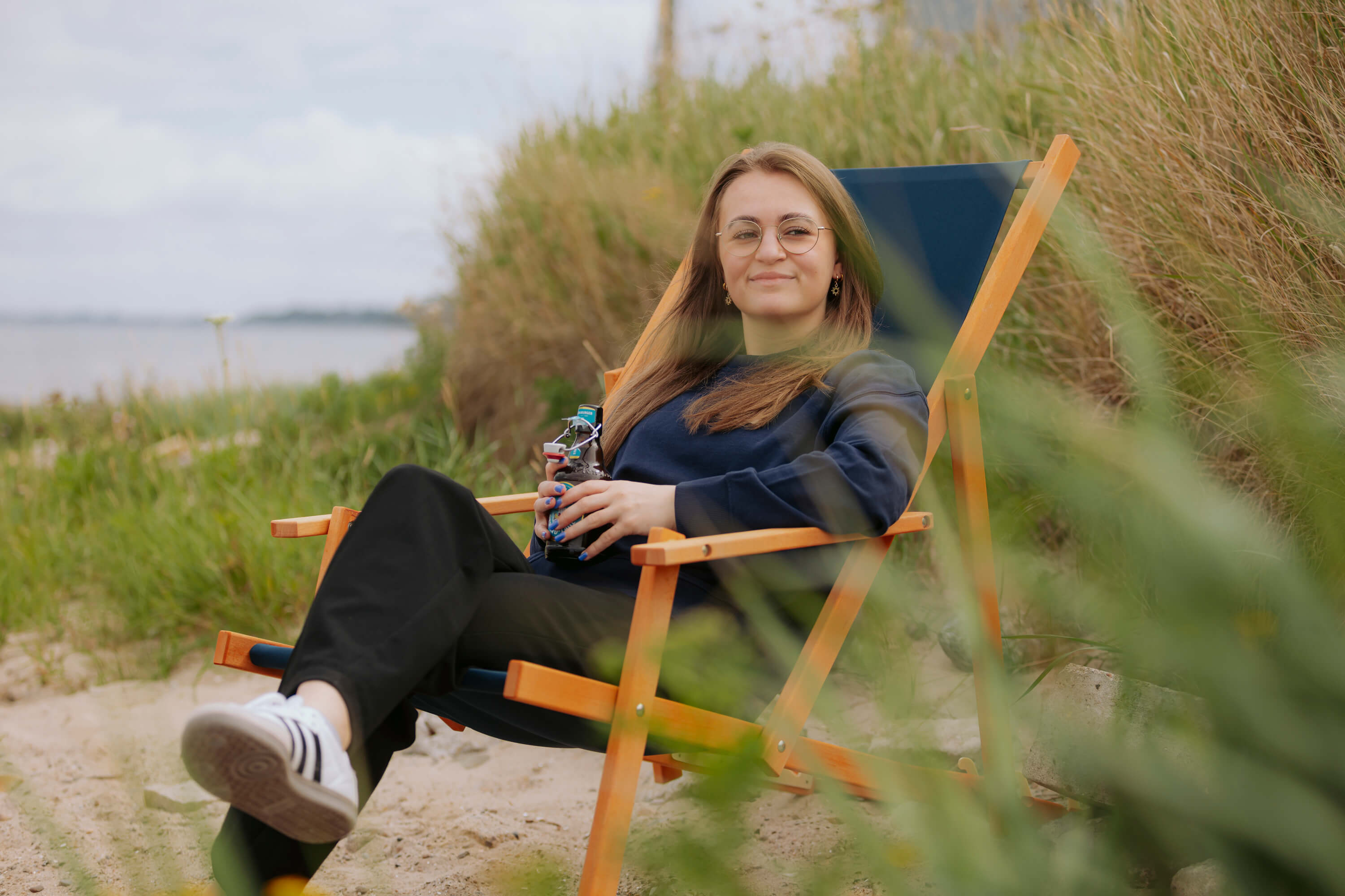 Eine Frau sitzt entspannt in einem blauen Liegestuhl aus Buchenholz am Strand und hält eine Flasche in der Hand. Im Hintergrund ist Strandgras zu sehen.